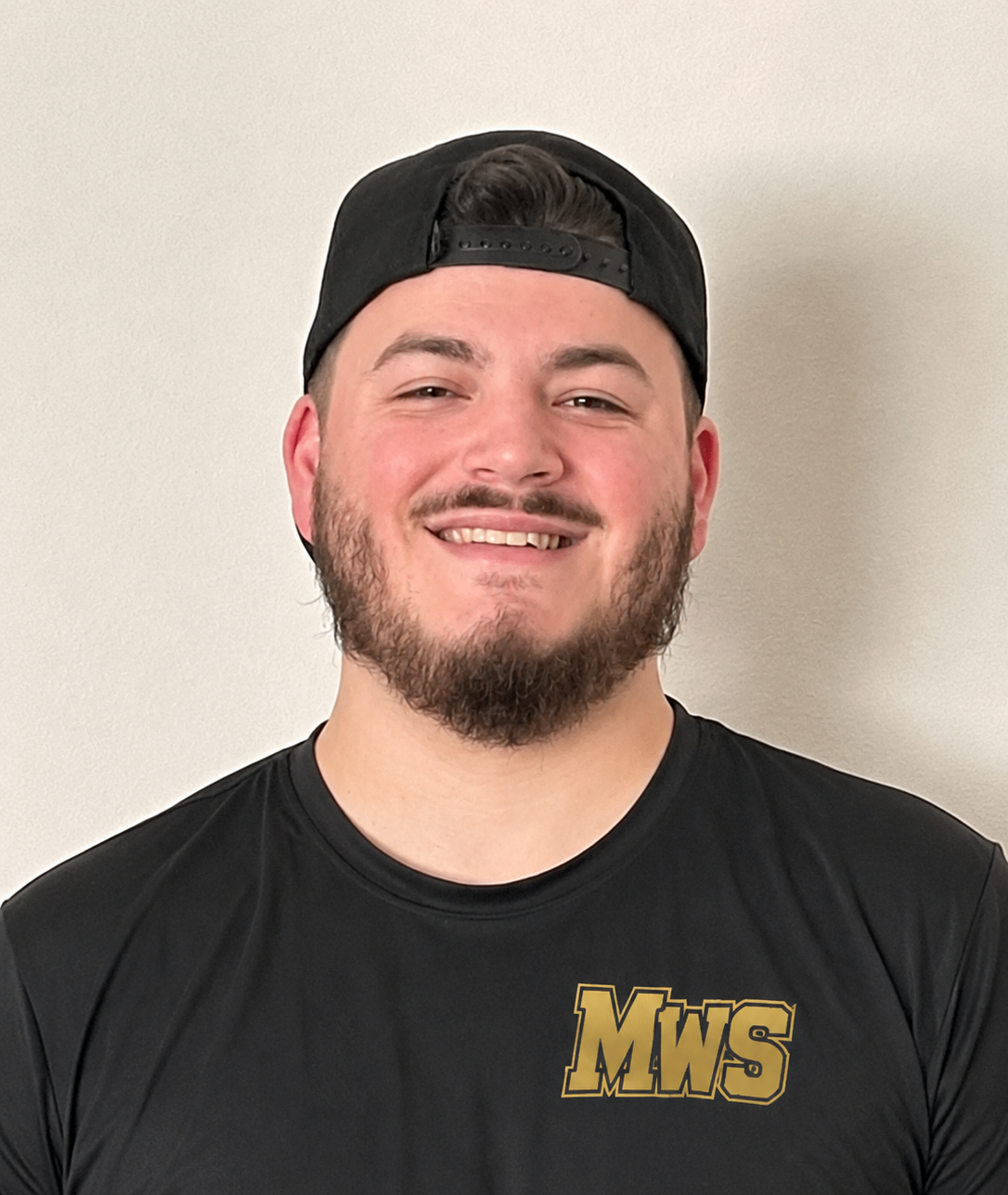 A smiling young man with a beard, wearing a black baseball cap backwards and a black t-shirt with gold lettering 'MWS', standing against a plain beige wall.