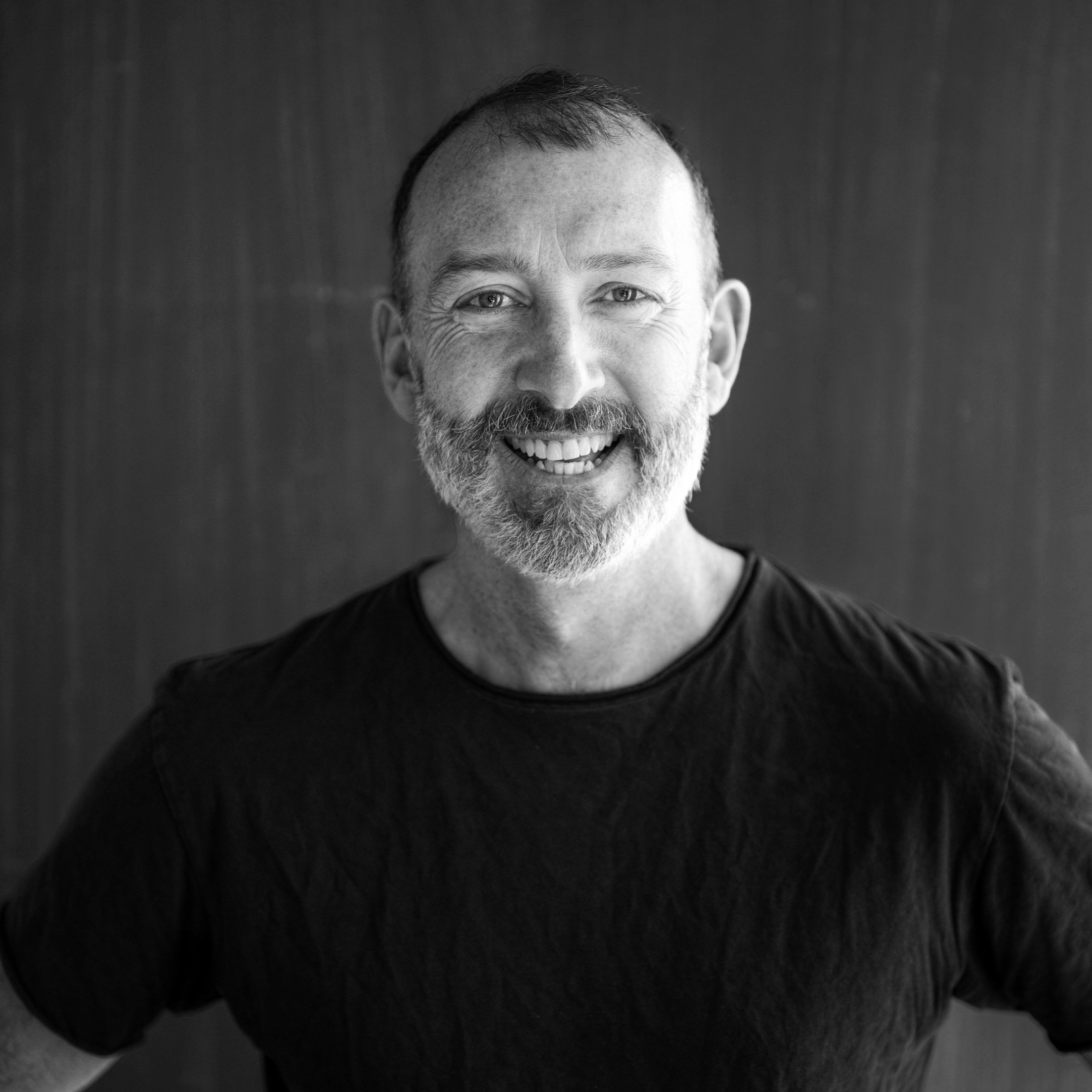 Black and white portrait of a smiling man with a beard, mustache, and short hair, wearing a black t-shirt, standing in front of a plain background.