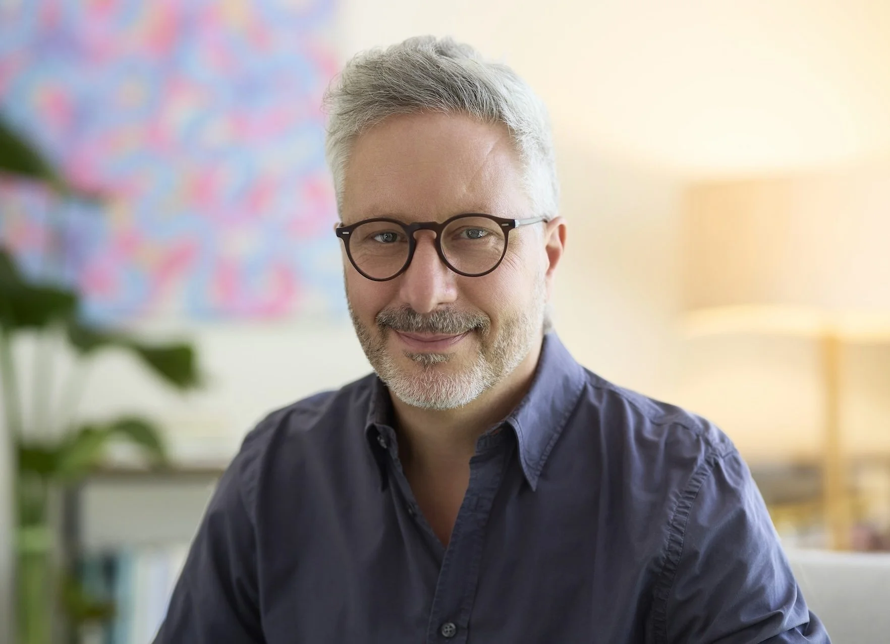 A middle-aged man with gray hair and beard, wearing black-rimmed glasses and a navy blue button-up shirt, smiling in an office setting.
