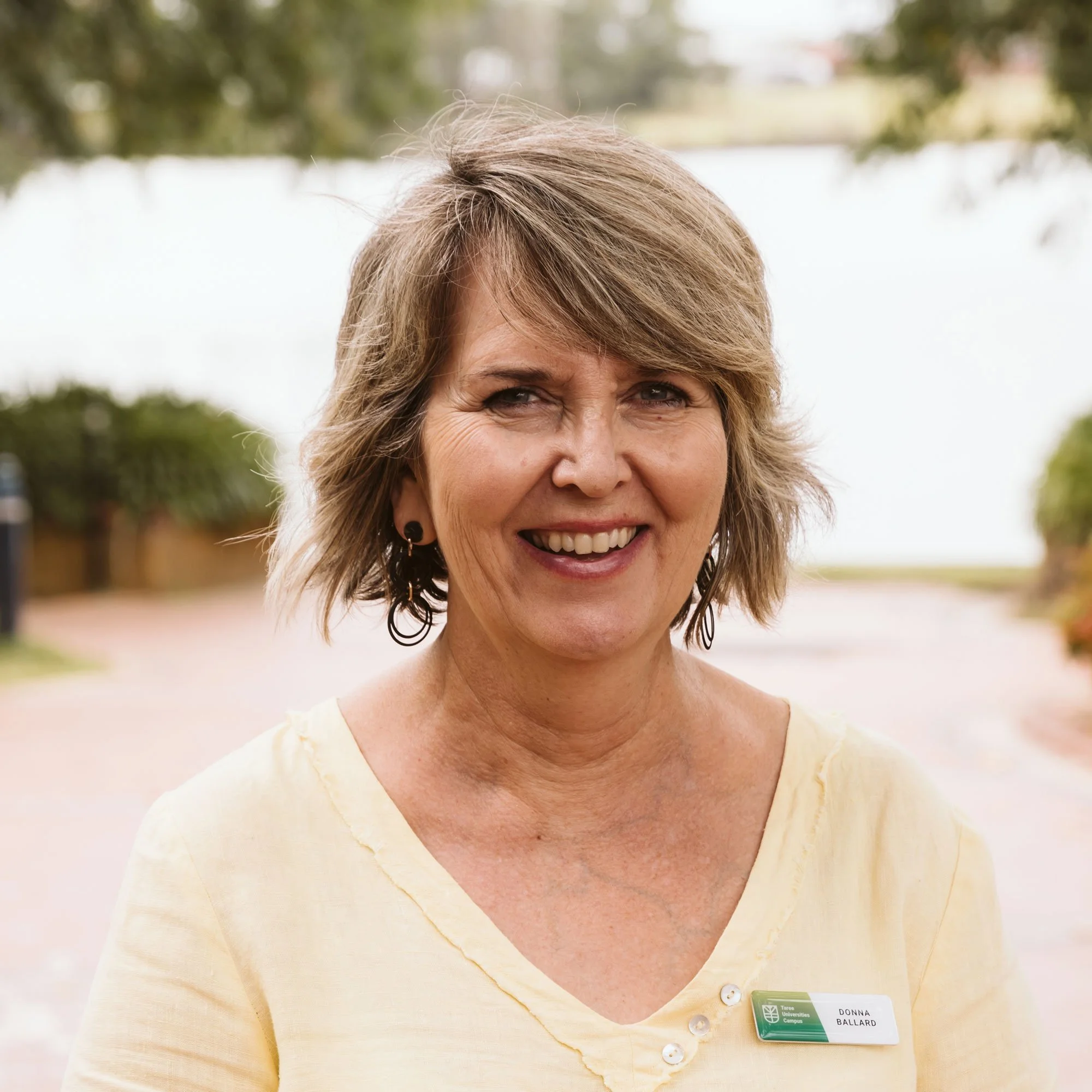 Smiling woman wearing a yellow shirt and name tag, outdoors with trees and a path in the background.