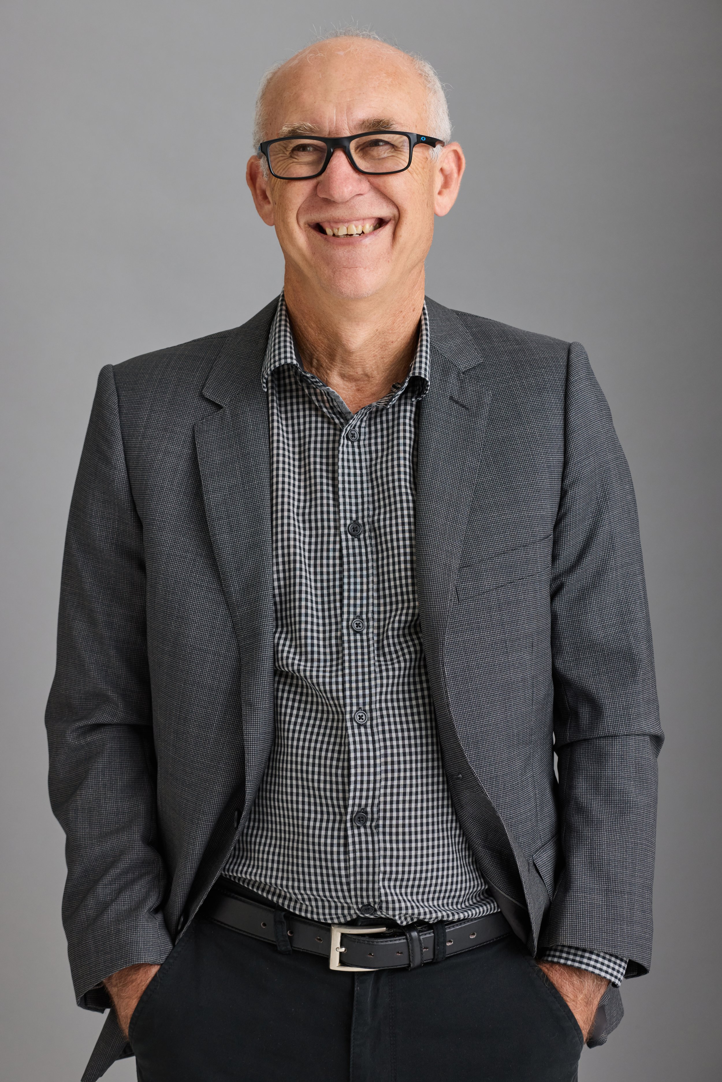 Smiling professional coach in glasses wearing a suit jacket and checkered shirt against a neutral background.