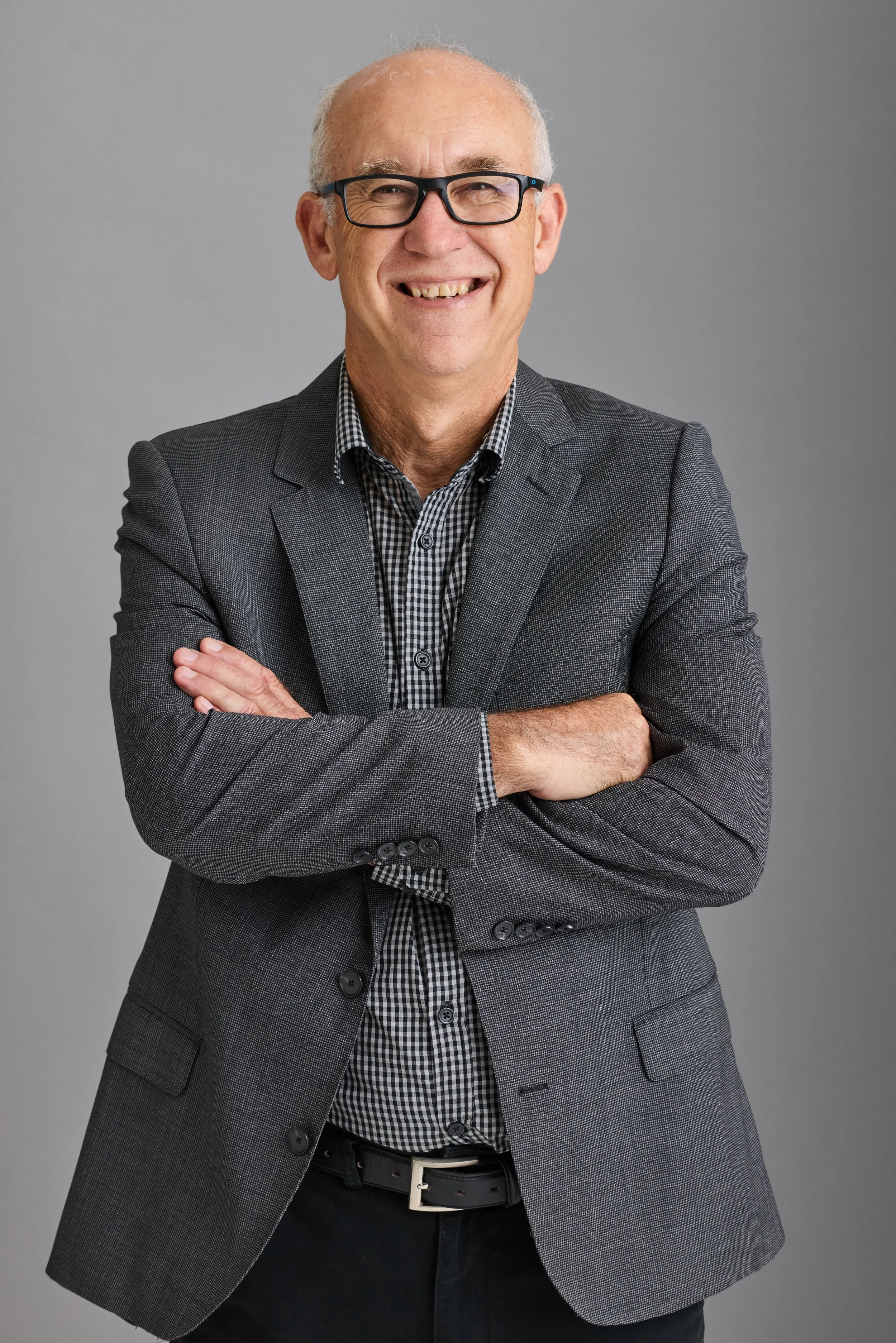 Smiling man in glasses wearing a gray blazer and checkered shirt, arms crossed, against a neutral background.