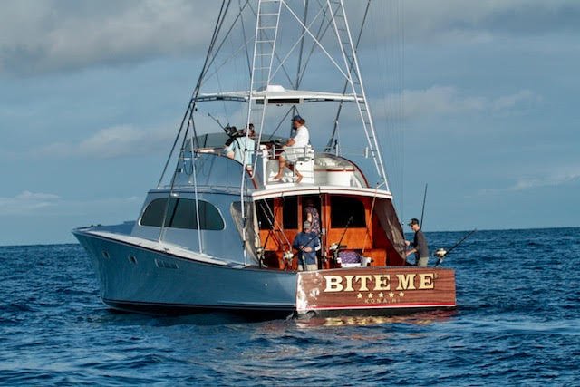 A sailboat named 'BITE ME' with people on board fishing in open water.