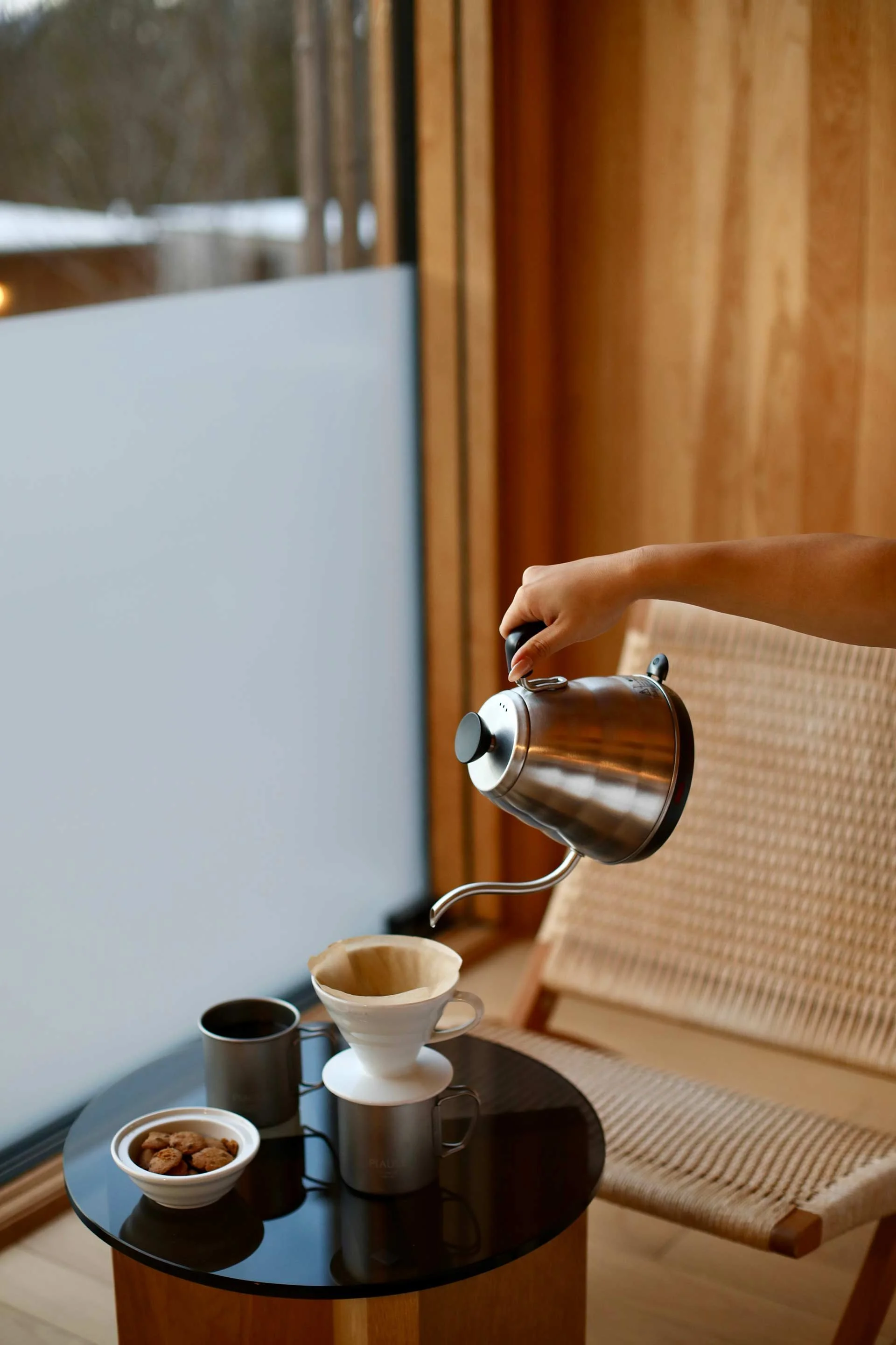 Person pouring hot water through a coffee drip cone placed on a mug, with additional mugs and a small bowl of cookies on a black round table in front of a chair.