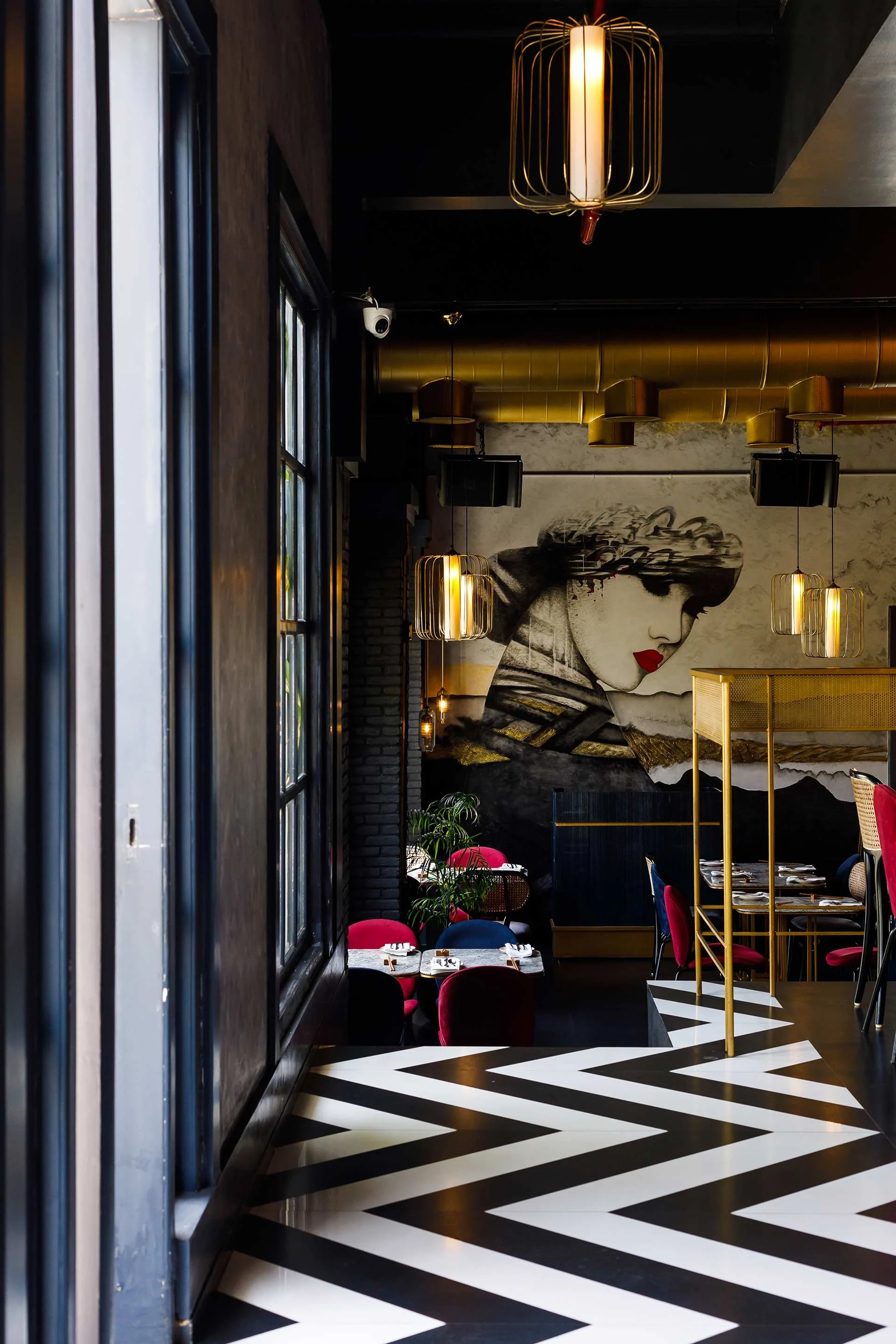 Interior of a stylish restaurant with black and white chevron flooring, a large wall mural of a woman with red lips, modern pendant lights, and red and blue velvet chairs.