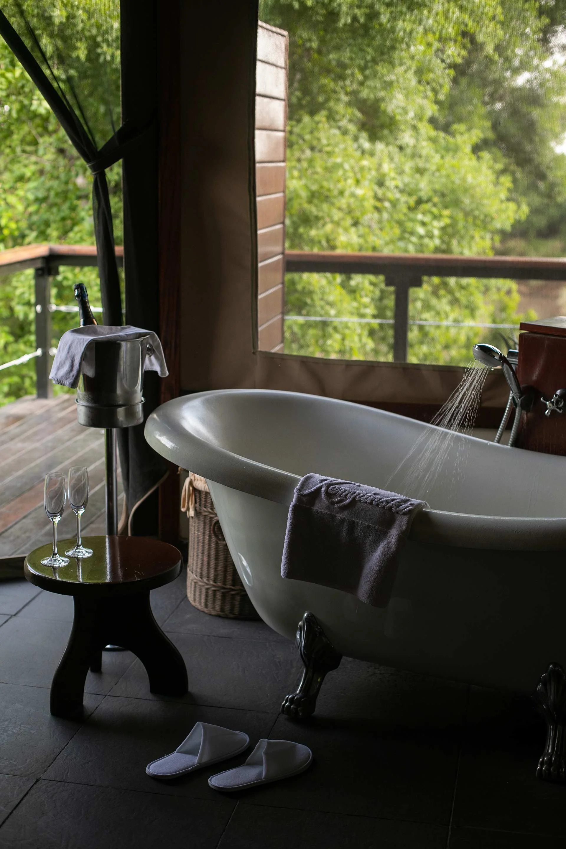 A rustic bathroom with a clawfoot bathtub, a small wooden stool with two champagne glasses, a bucket with a towel and a bottle, slippers on the floor, and a view of green trees. At Olare Mara Kempinski, in Masai Mara.