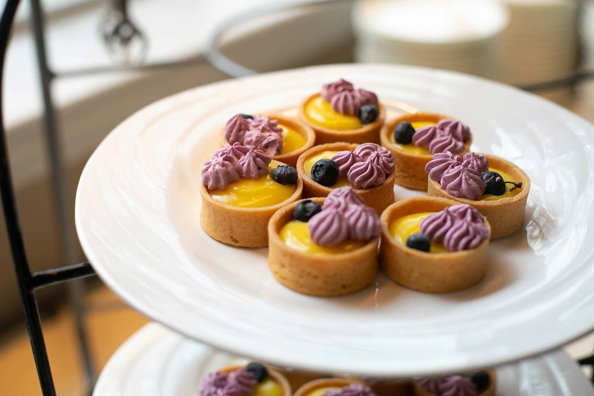 A white serving plate with small lemon tartlets topped with purple piped cream and blueberries.