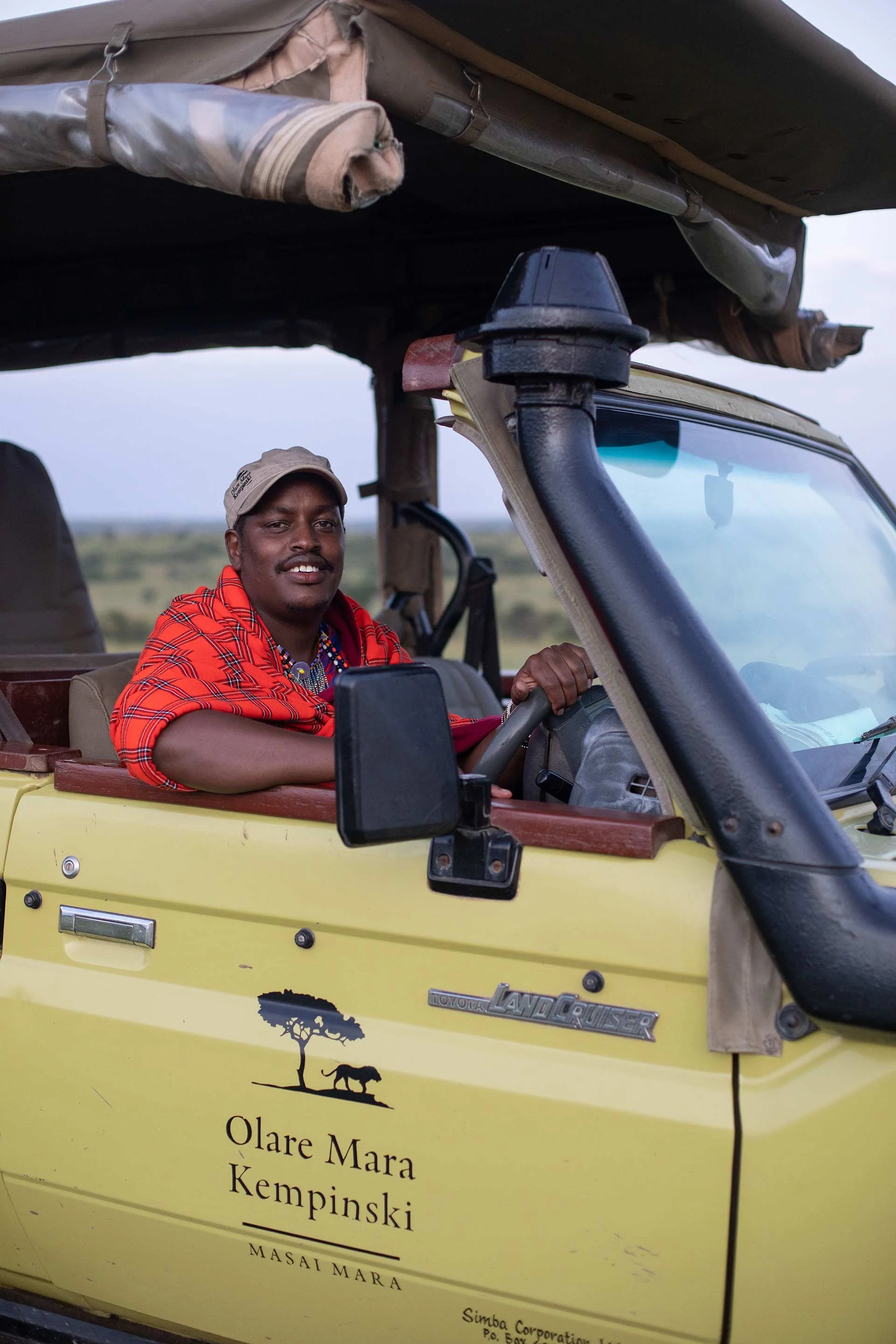 A man sitting in the driver's seat of a yellow safari vehicle with a canopy, smiling. The vehicle has a logo and text on the side that reads 'Olare Mara Kempinski' and features a logo of a tree and an animal. The background shows an open landscape.