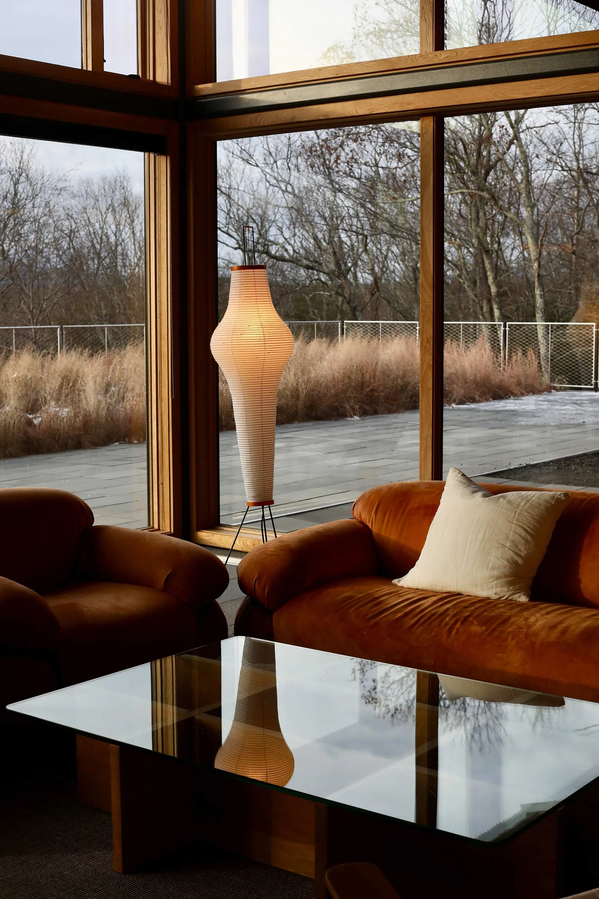 A cozy living room with large floor-to-ceiling windows showing an outdoor deck and leafless trees, furnished with a brown armchair, a matching sofa with a cream pillow, a glass coffee table, and a tall, curved white floor lamp.