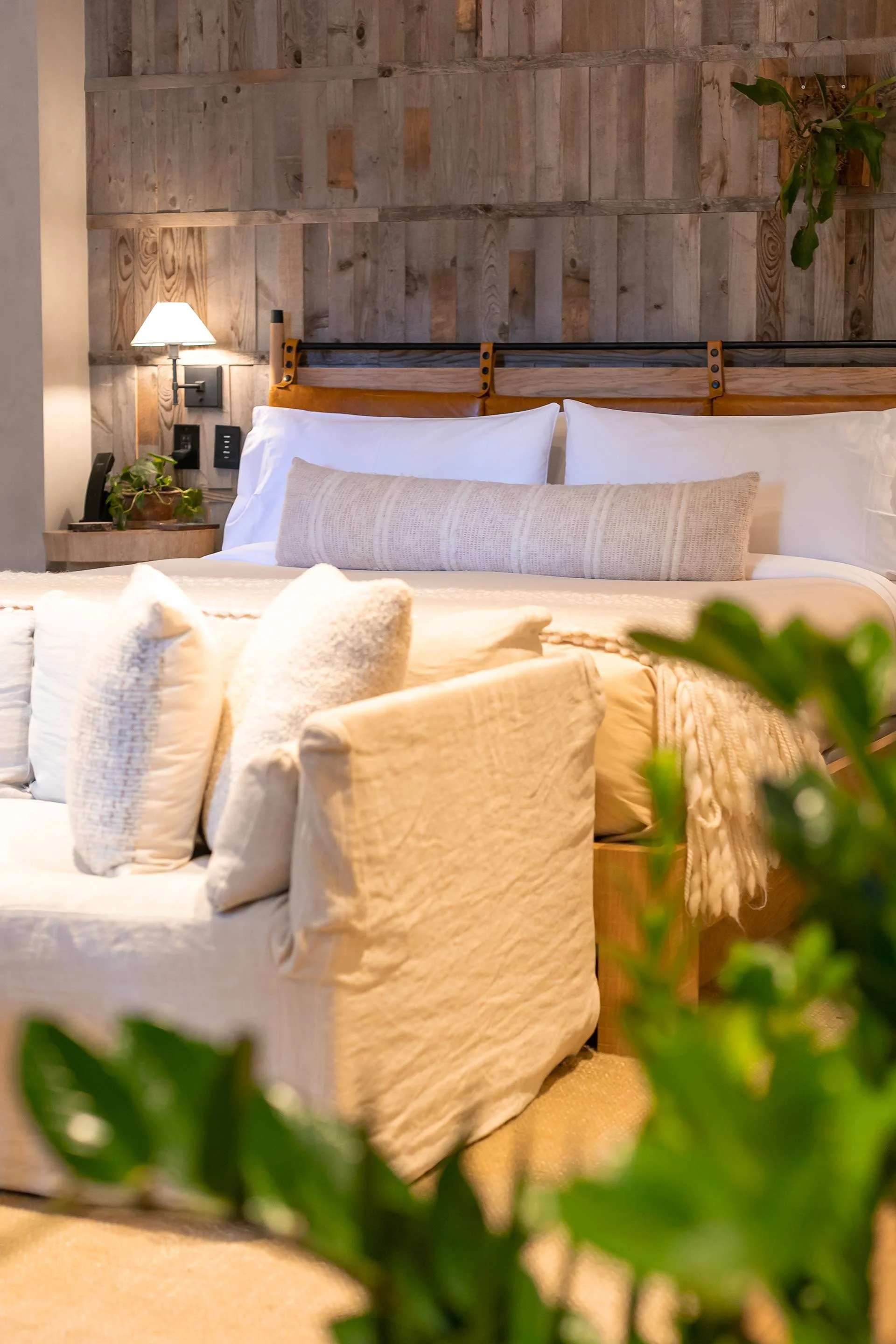A cozy hotel bedroom with a wooden headboard, white pillows, a beige bolster pillow, a bedside lamp, and some greenery.