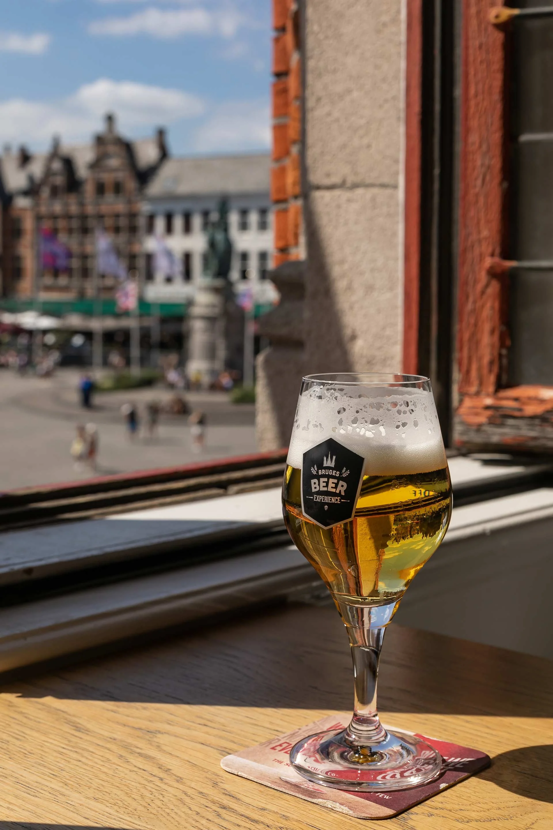 A glass of beer with a foam head sitting on a coaster on a wooden table near a window, looking out onto a city square with historic buildings and flags.