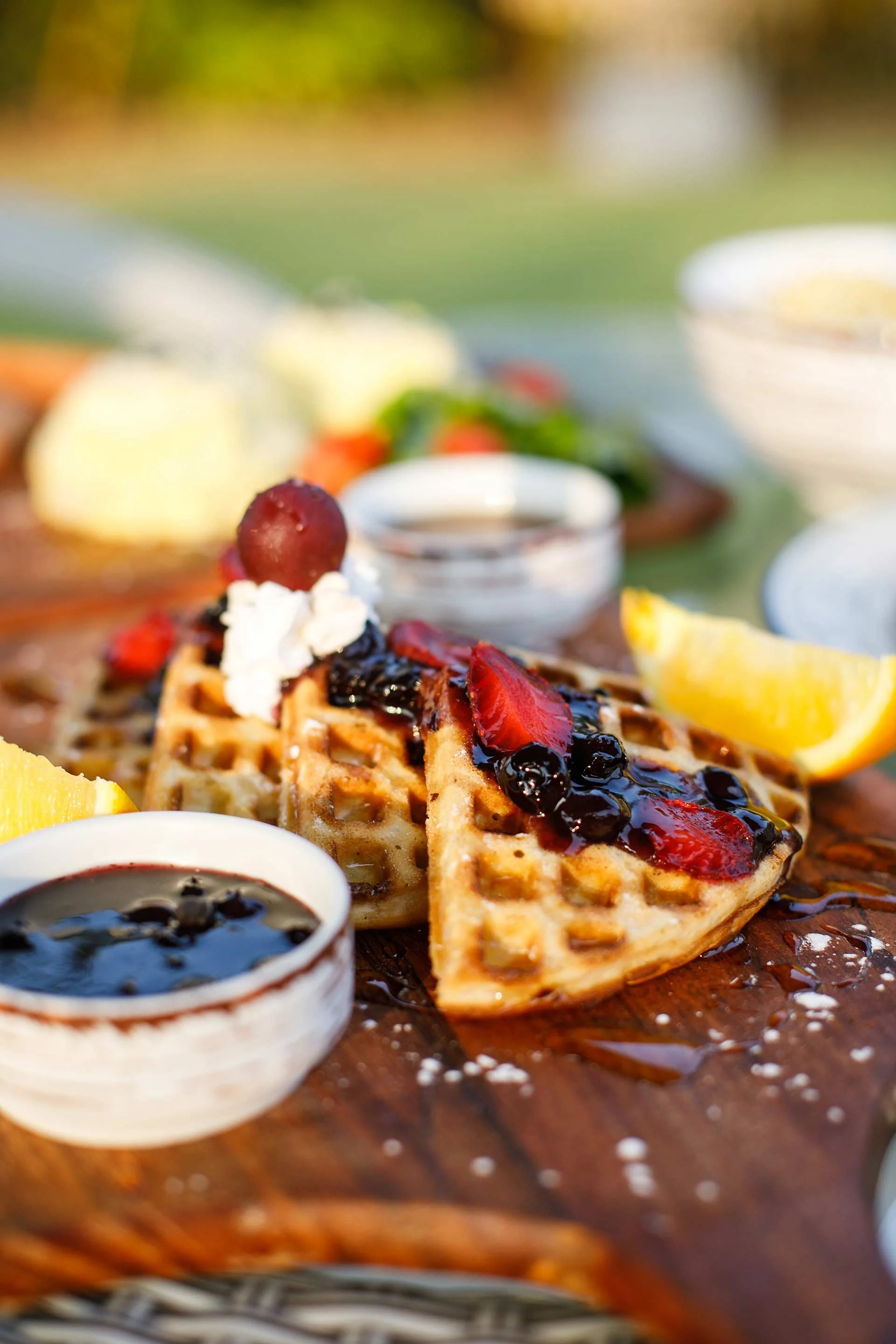 A breakfast scene with waffle topped with strawberries, blueberries, whipped cream, and syrup, served with lemon wedges and extra syrup in small bowls on a wooden table outside.