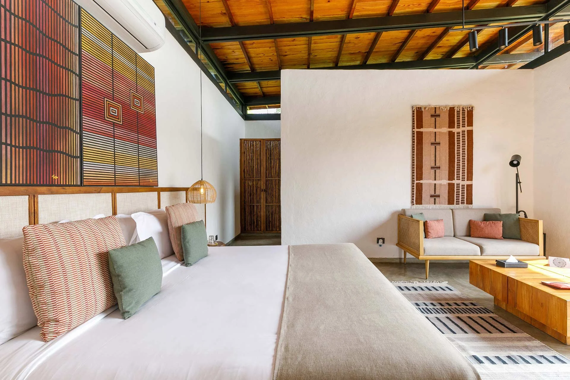 A modern bedroom with a large bed, multiple pillows, and a bedside lamp. There is a sitting area with a sofa, patterned wall hanging, and wooden furniture in a room with a high, wooden ceiling.