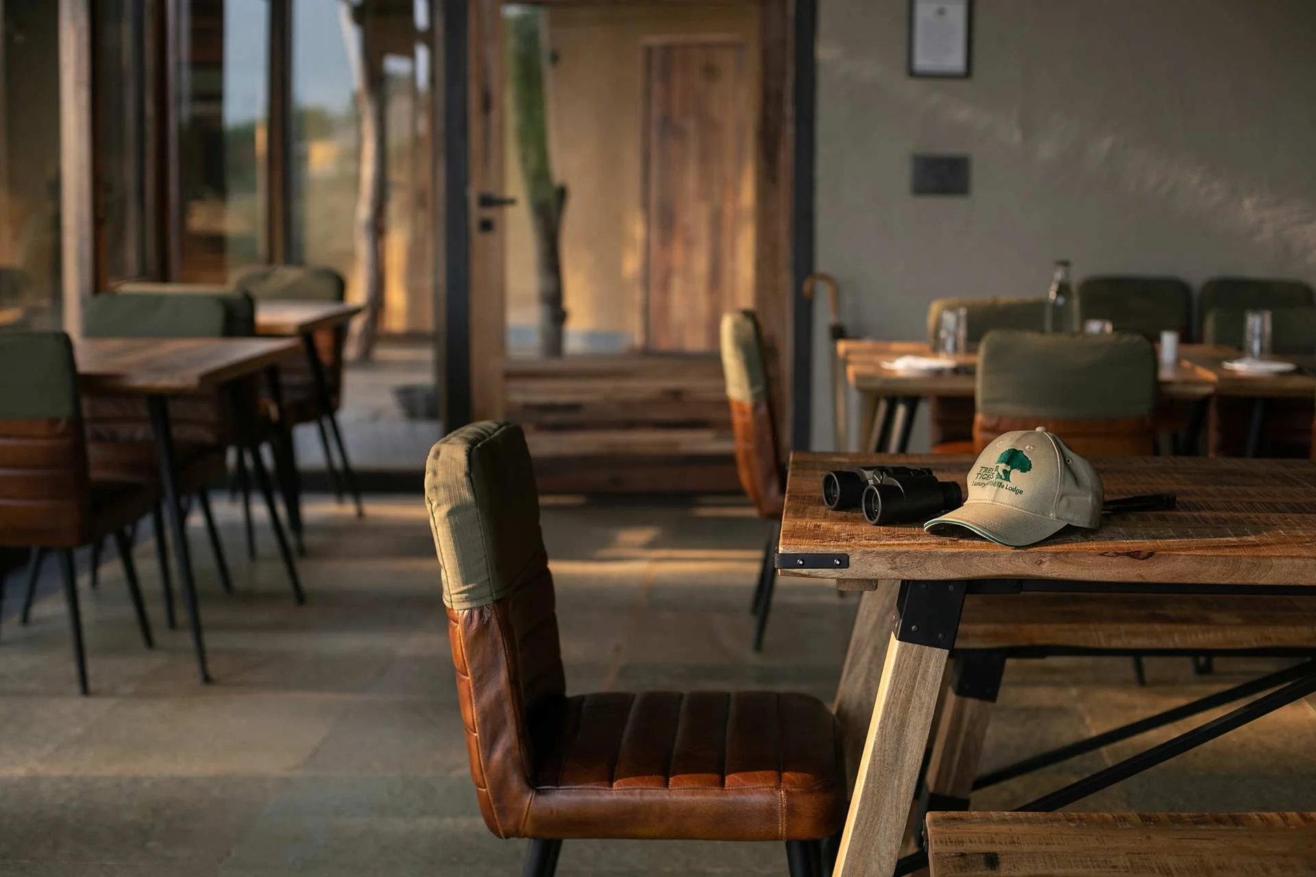 Empty rustic restaurant with wooden tables and green cushioned chairs, binoculars and a hat on one table, large windows, and a wooden door.