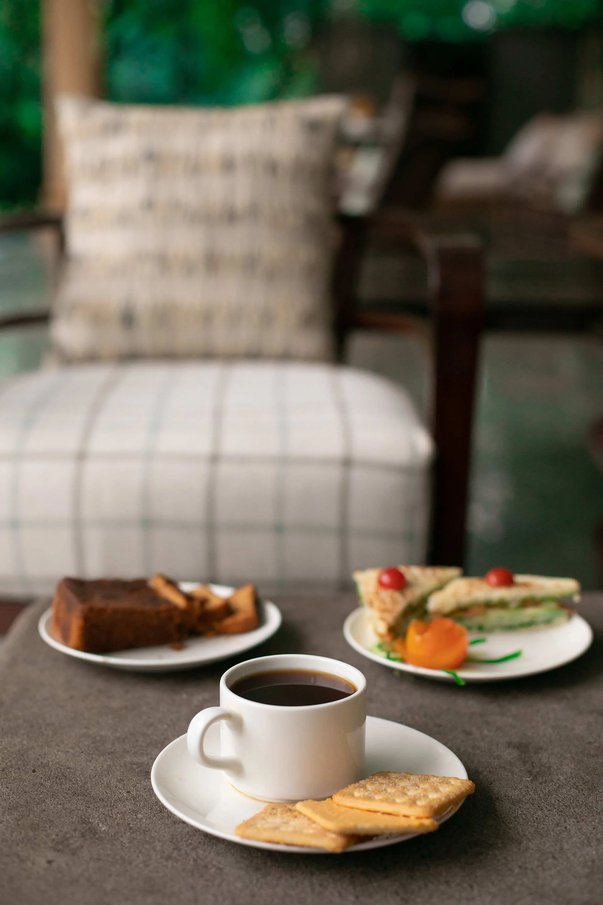 A cup of black coffee on a saucer with a few biscuits, a slice of chocolate cake, and a sandwich with cherry tomatoes, lettuce, and a peach on a plate, set on a table with a blurred cozy chair and sofa in the background.