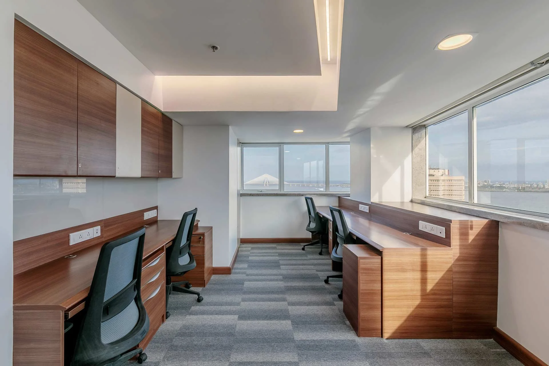 An empty office space with four work desks with black office chairs, large windows with city and water views, wood cabinets, and electrical outlets.