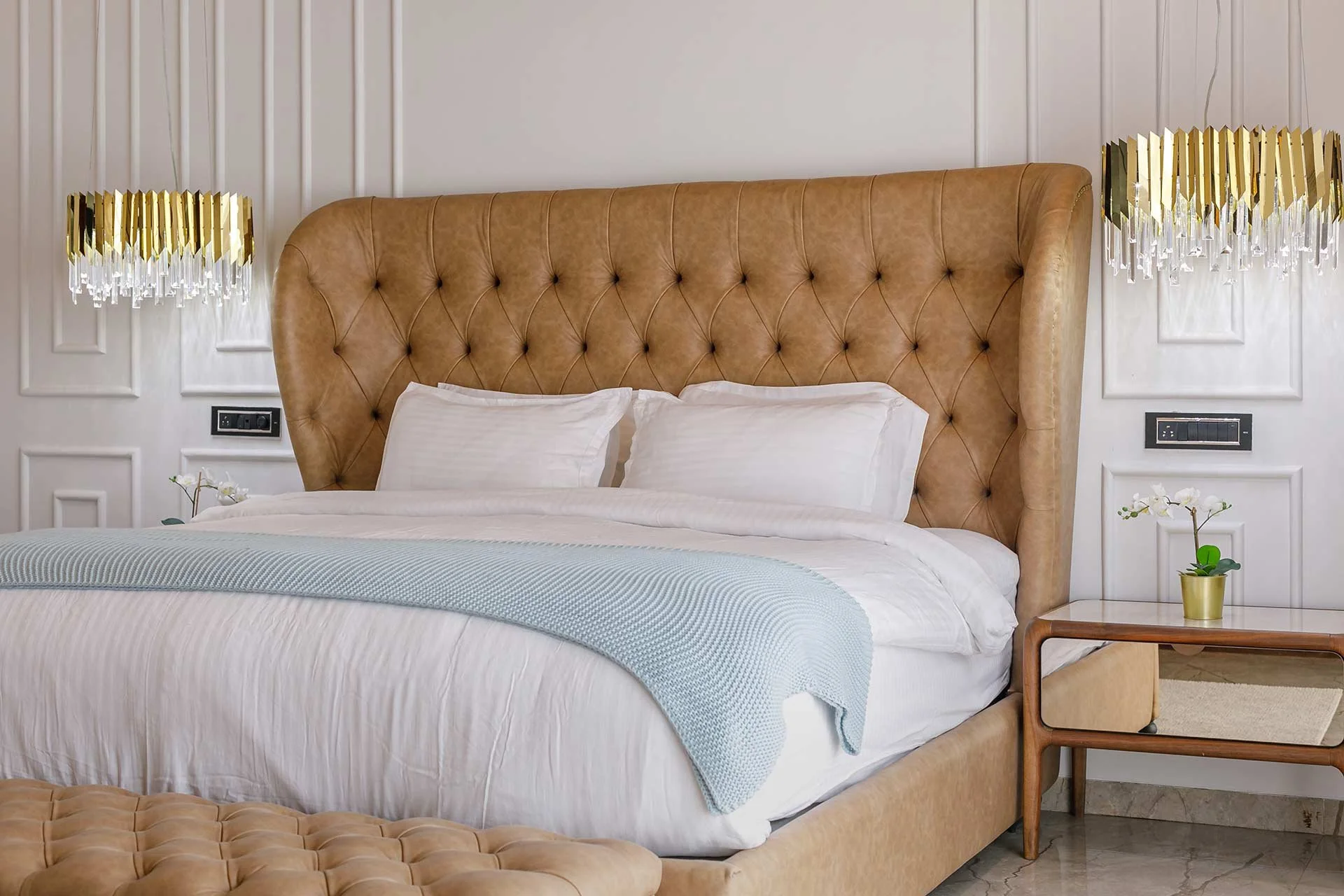 A bedroom with a large tan tufted headboard, white bedding, and pillows. There are two gold chandeliers with crystal accents on either side. A small wooden nightstand with a plant in a gold pot is beside the bed.