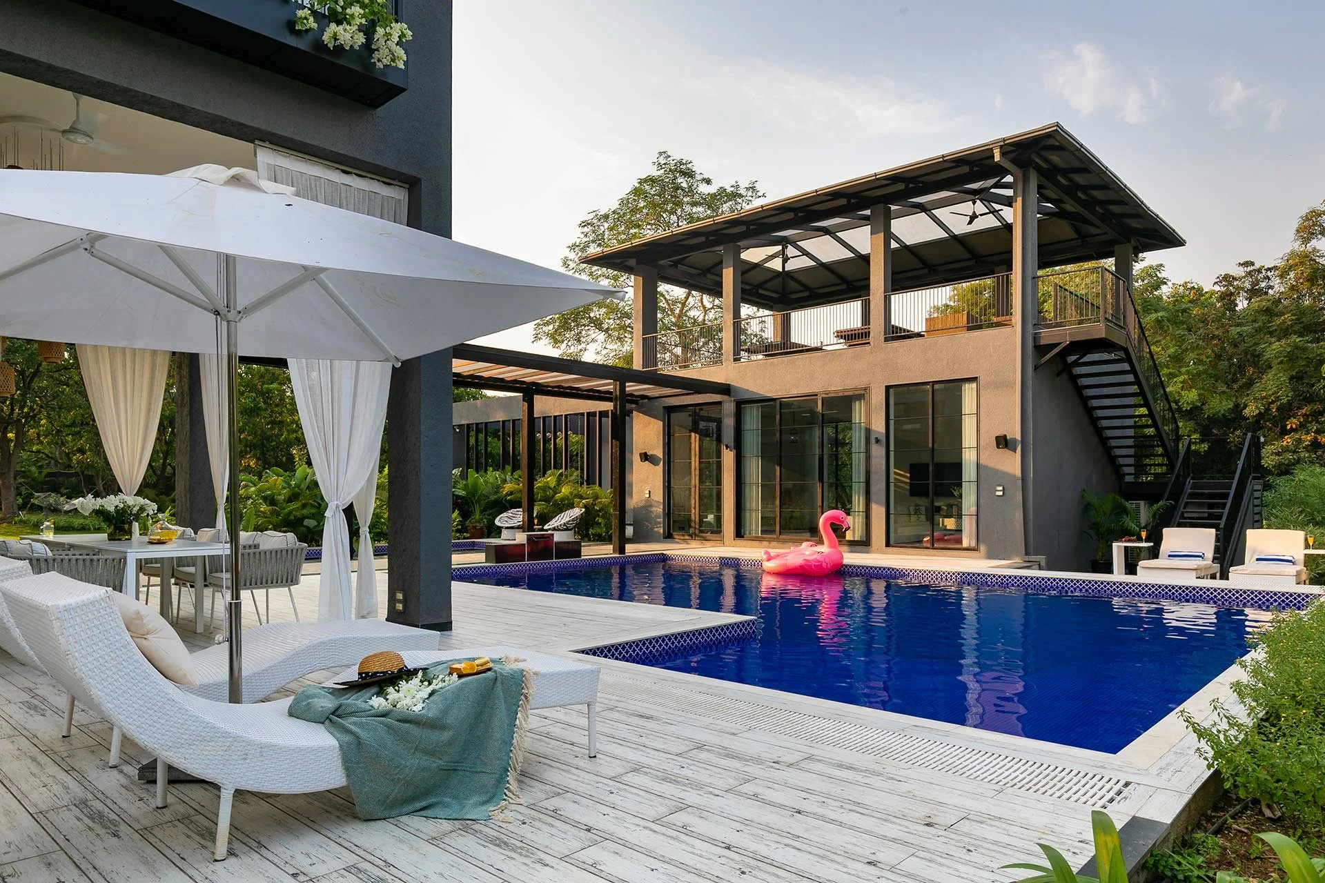 Modern house with a pool, inflatable pink flamingo, outdoor furniture, and large white parasol. In Alibuag, at Charcoal villa, photographed by Rishul Bangar.