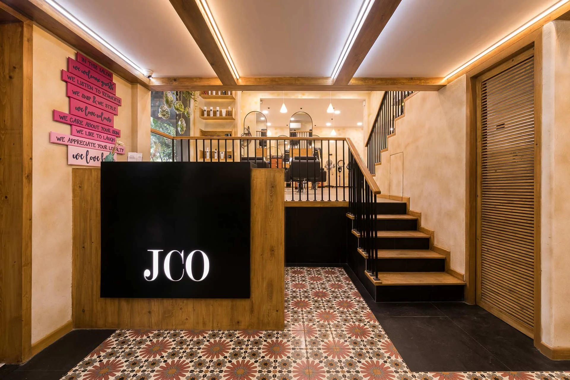 Interior of a modern beauty salon with a reception desk displaying the initials 'JCO' in white on a black background. The salon features wooden accents, patterned tile flooring, and a staircase leading to a seating area with black chairs and mirrors.