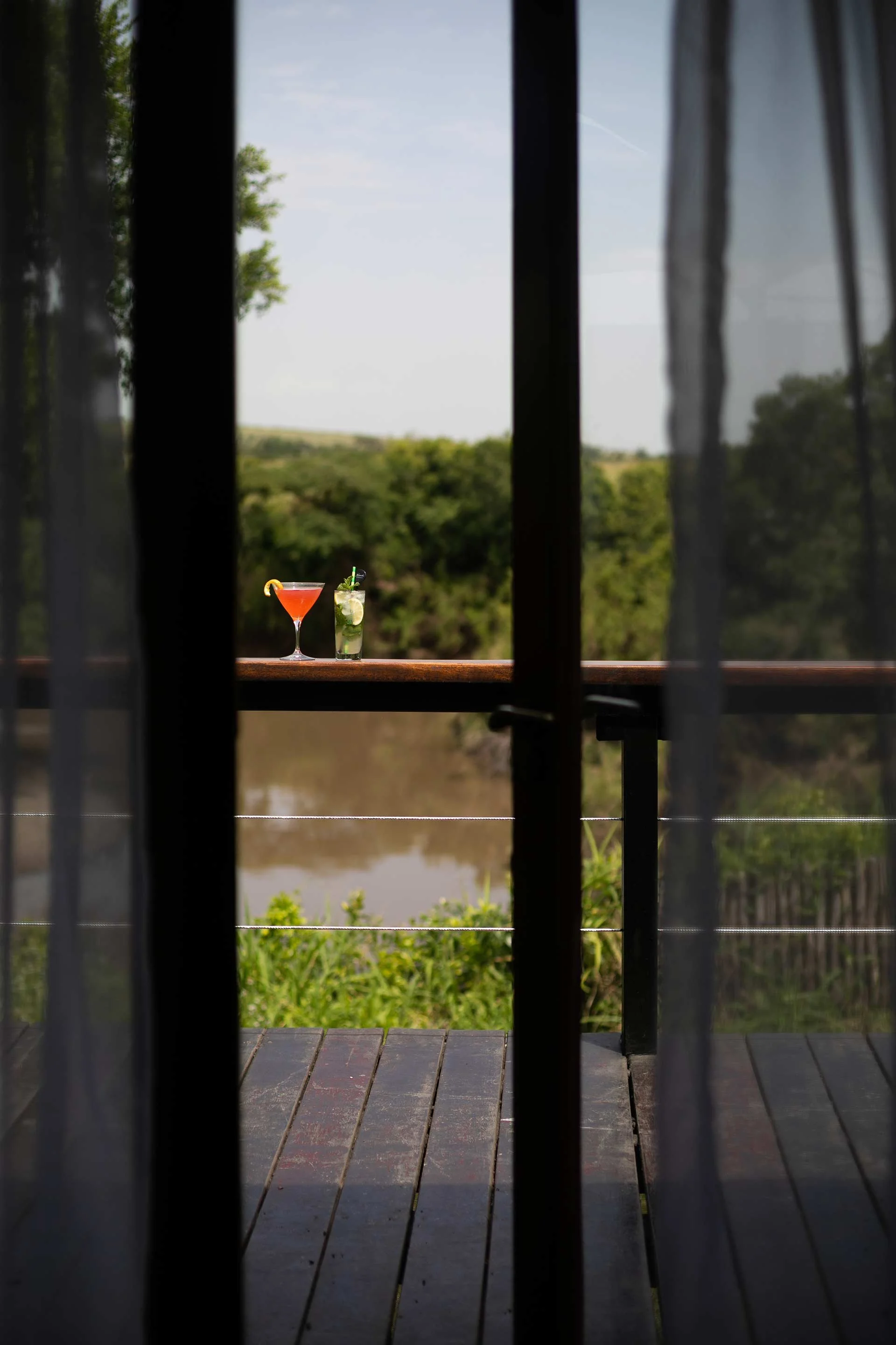 Two cocktails, one red and one green, on a wooden railing of a balcony overlooking a river and green trees.