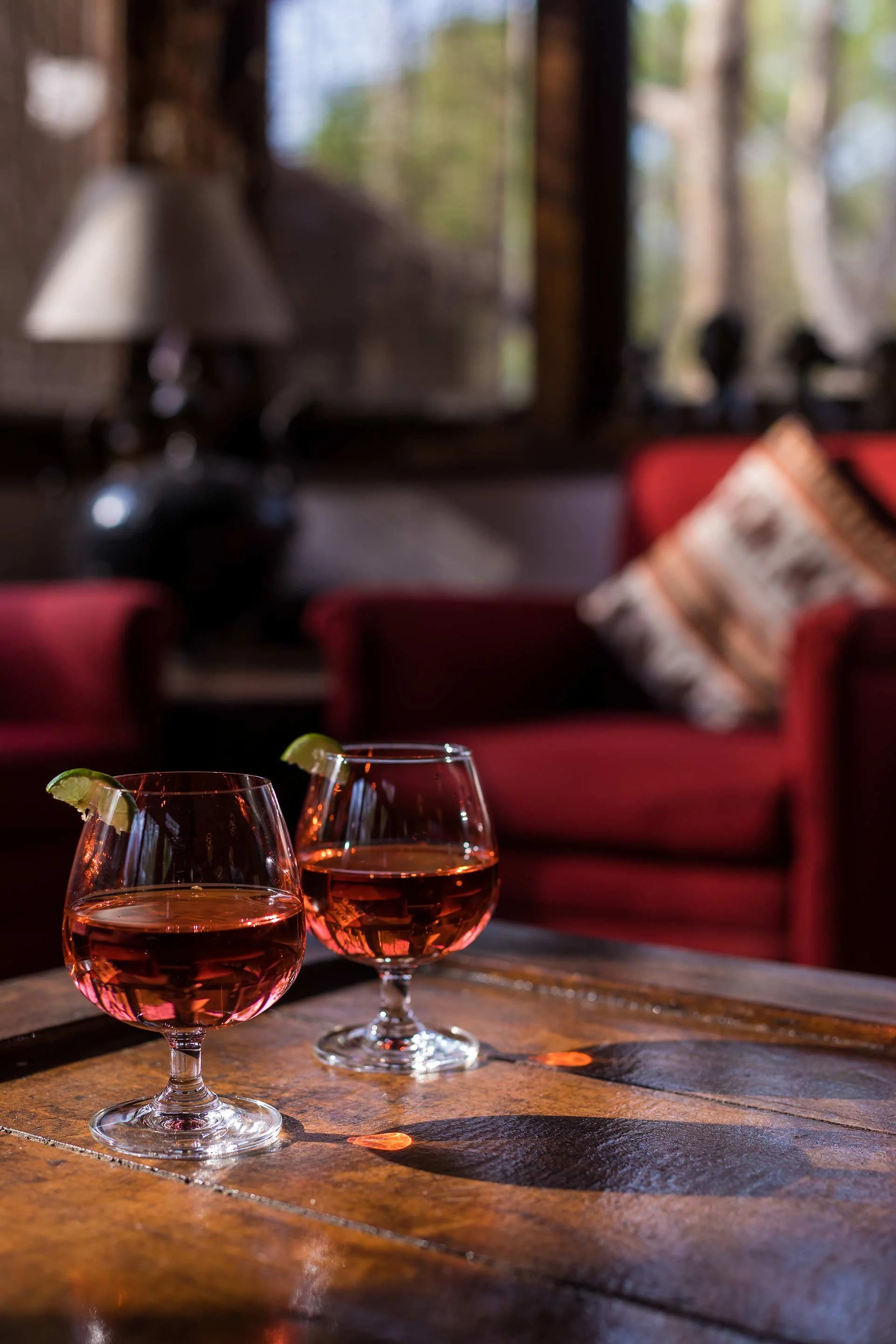 Two glasses of pink cocktail with lime wedges on a wooden table inside a cozy living room with red sofas and a large window in the background.