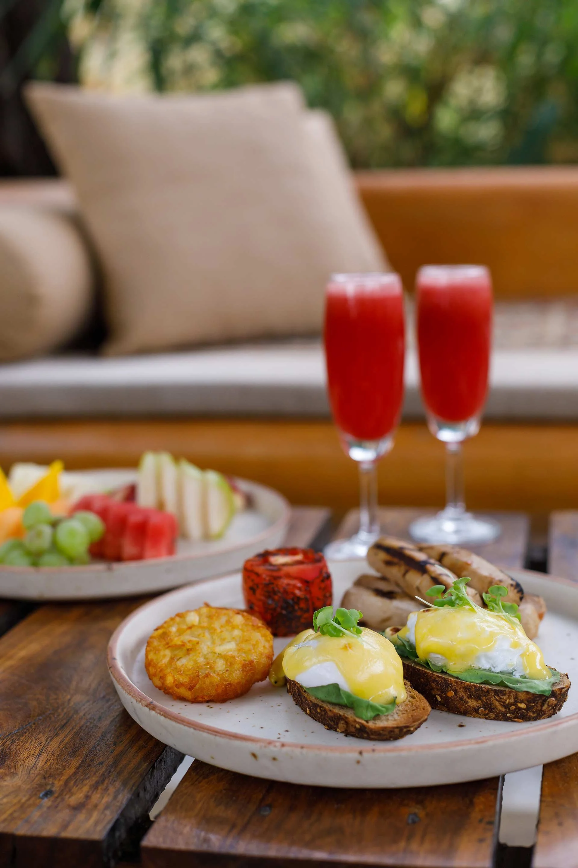 A breakfast plate with eggs Benedict topped with hollandaise sauce and microgreens, grilled bread with smoked sausage, a hash brown, and a red roll of salmon, with two glasses of red juice or smoothie in the background, and a fruit platter with grape