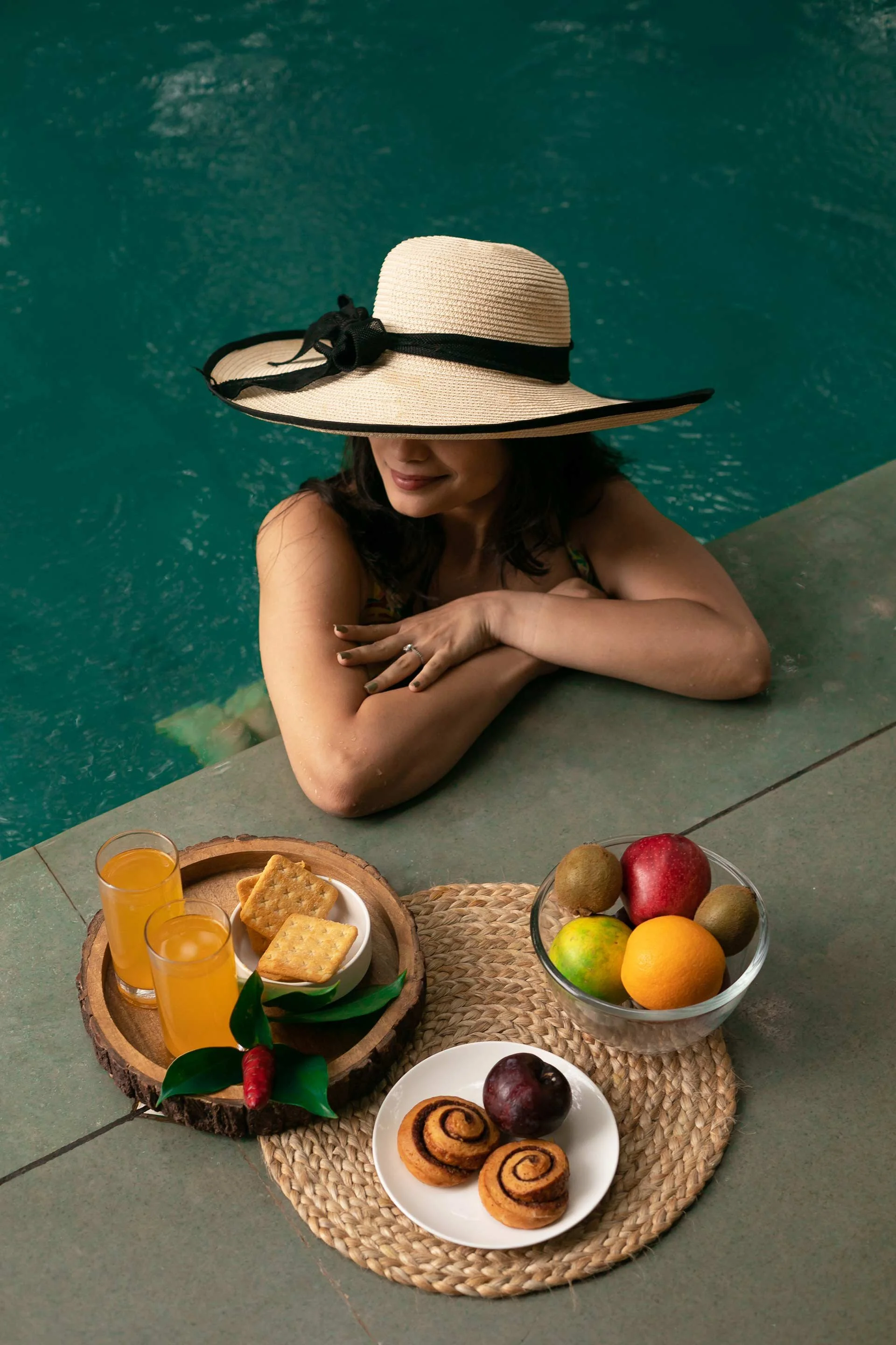 A woman leaning on the edge of a swimming pool, wearing a wide-brimmed straw hat with a black ribbon, with a table of snacks and fruits in front of her.