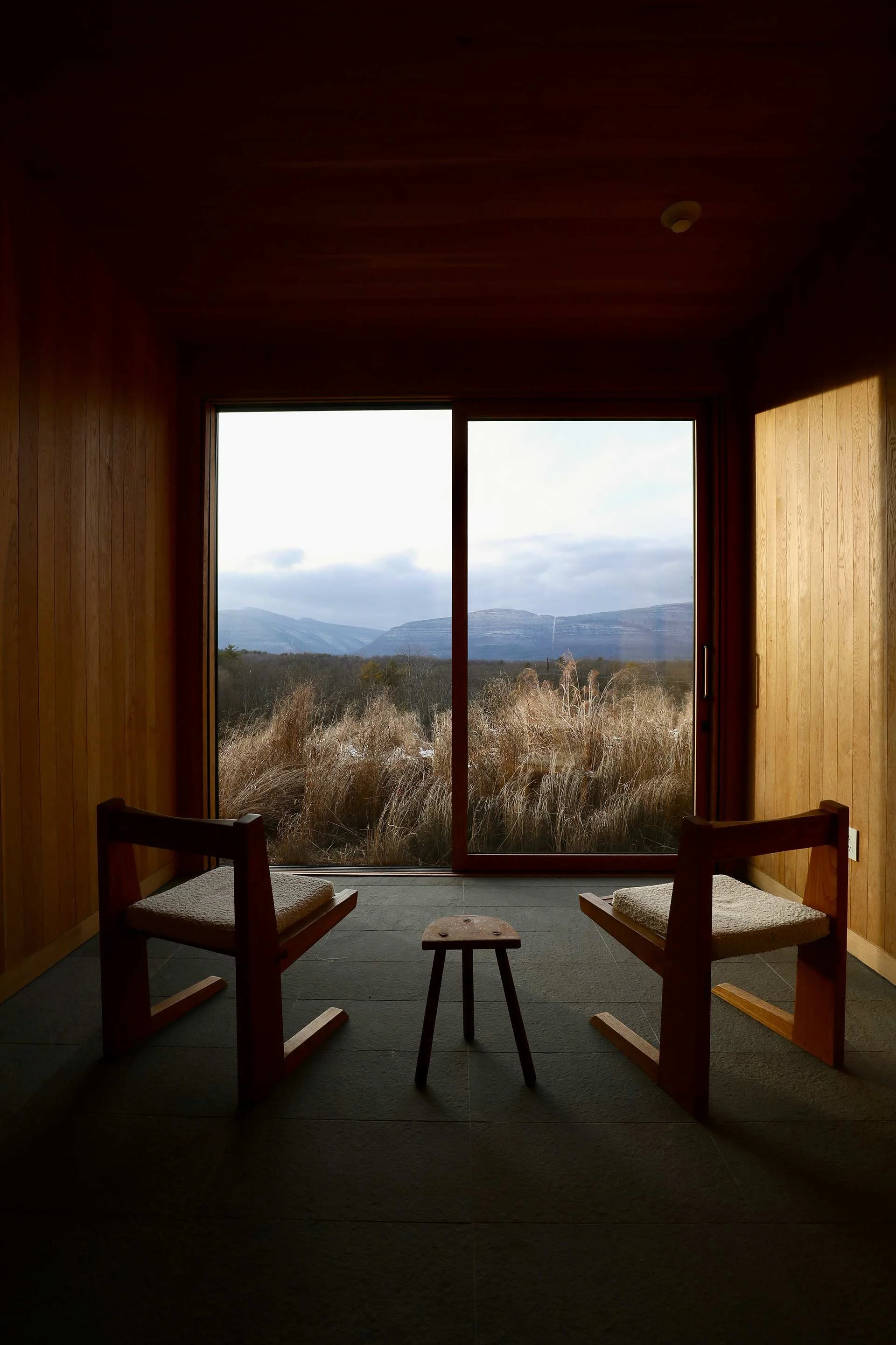 Interior of a room with wooden walls, two wooden chairs with cushions, a small wooden stool, and a large sliding glass door showing an outdoor landscape of grass, trees, mountains, and a cloudy sky.