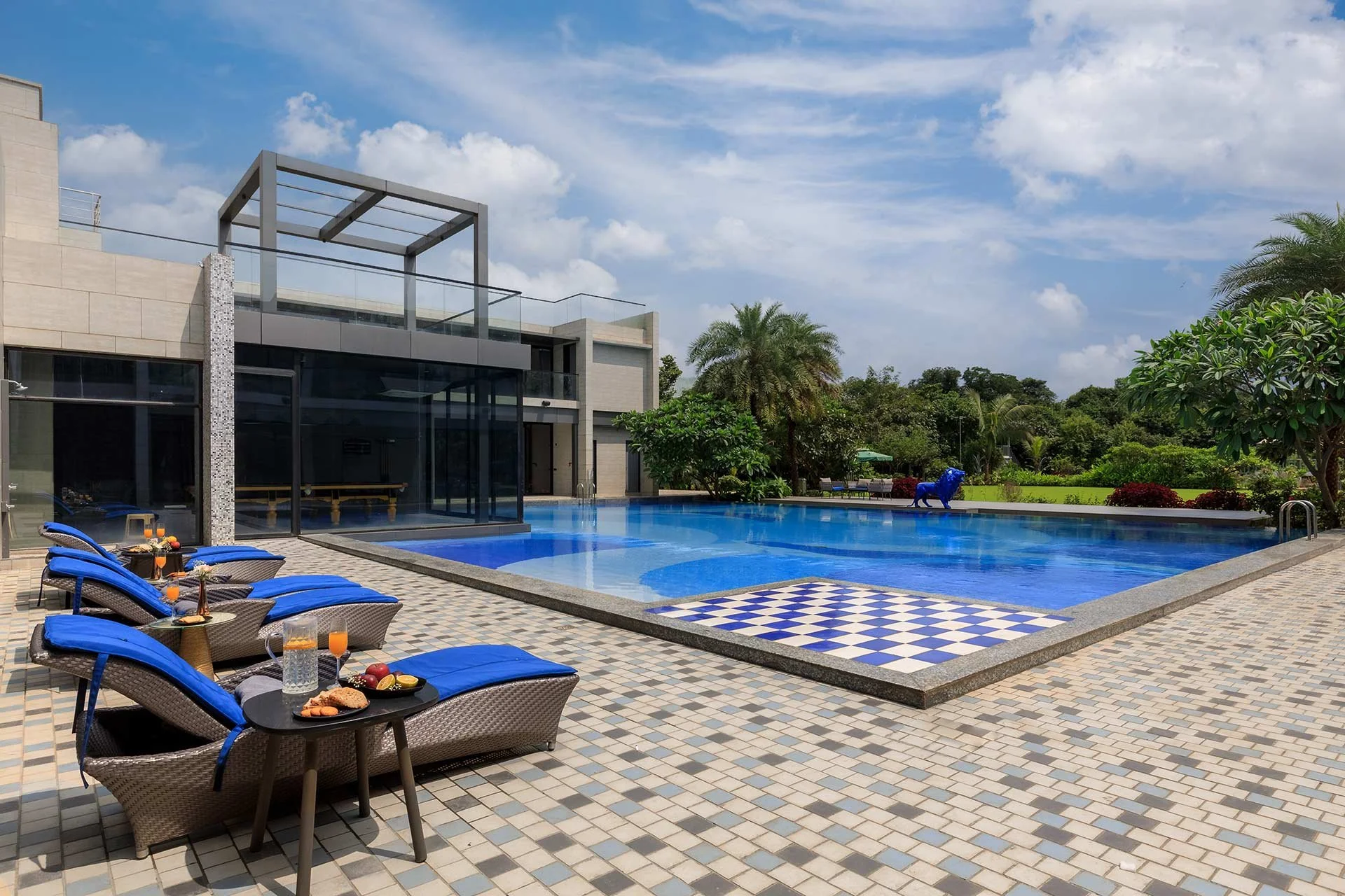 Luxury modern house with a swimming pool, lounge chairs, drinks, and a garden with trees under a blue sky. At Karjat, photographed by Rishul Bangar.