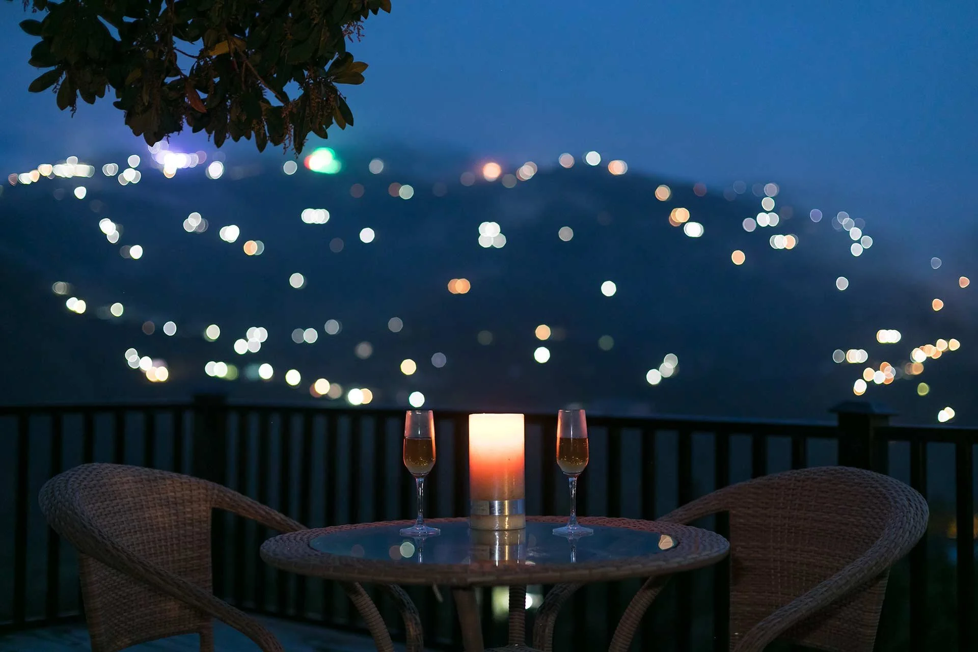 A nighttime scene of a balcony with a round glass table, two chairs, a lit candle in the center, and two glasses of champagne. The background is blurred with city lights on a hill.