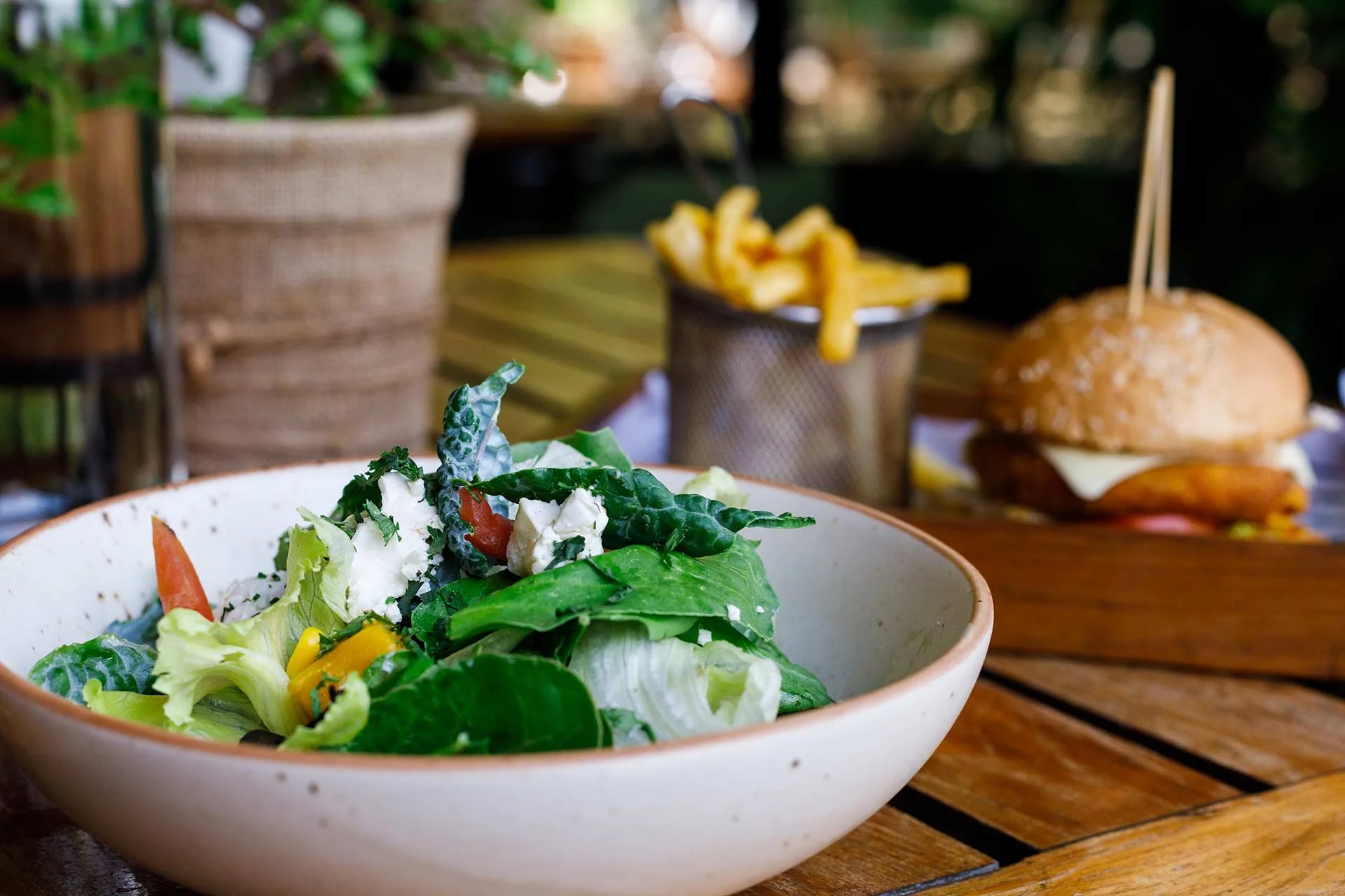 A bowl of fresh salad greens with vegetables and cheese in the foreground, with a cheeseburger and French fries in a metal container in the background, on a wooden table.