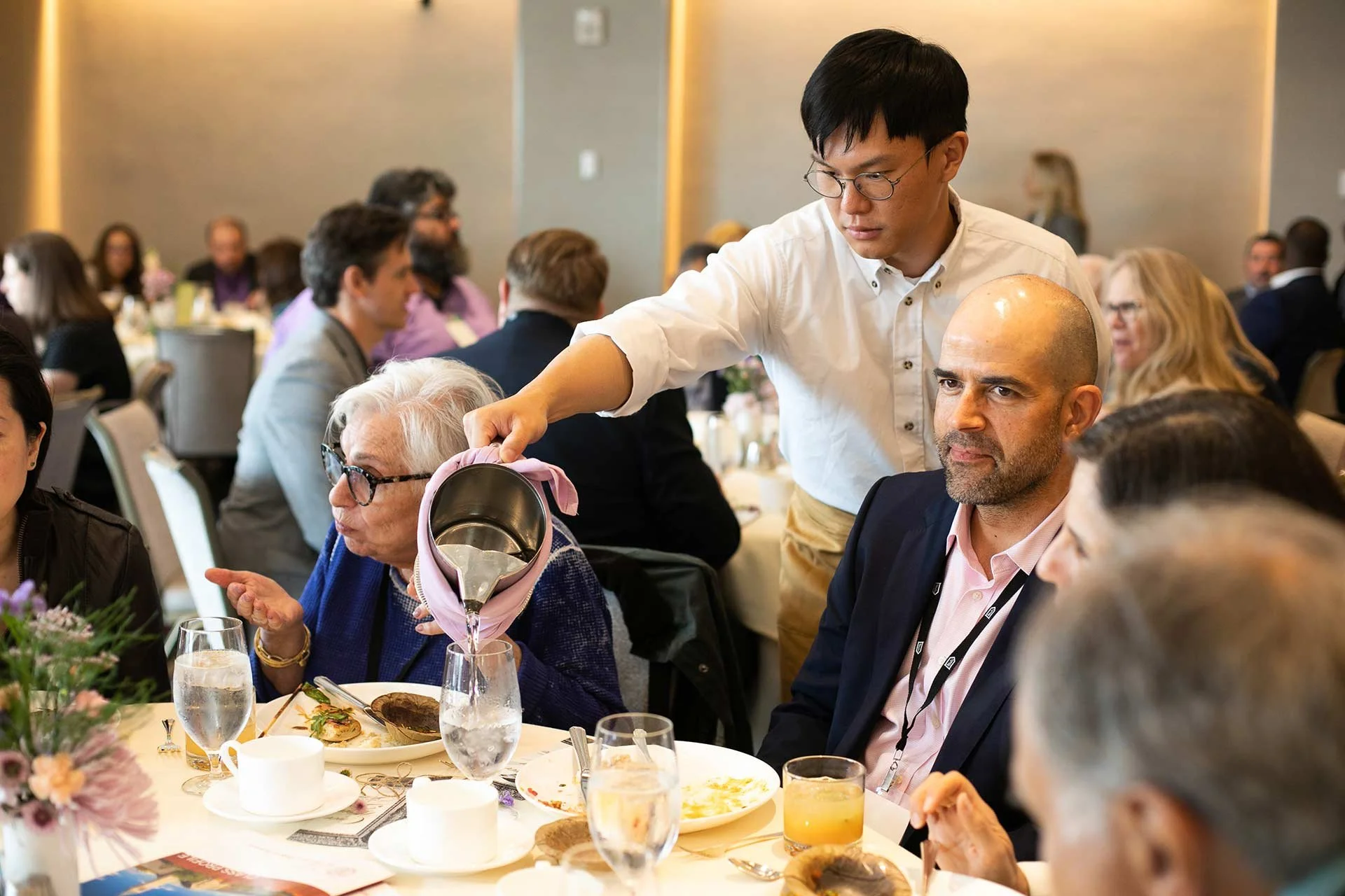 A man pours water from a pitcher into a woman's glass at a formal dinner event, surrounded by other guests.