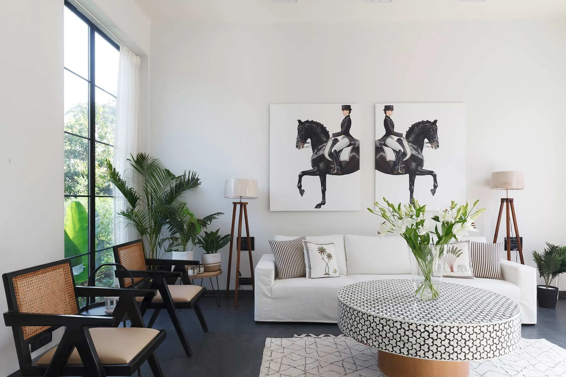 Living room with white sofa, black and white patterned coffee table, large glass window, houseplants, two black and white horse and rider artworks, and standing lamps.