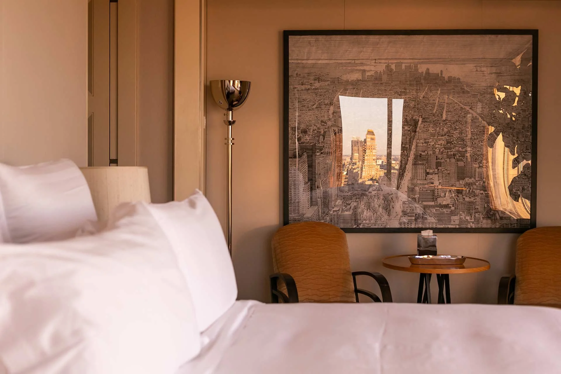 A hotel room with a bed, a framed cityscape photograph on the wall, two armchairs, a small round table, a tissue box, and a standing lamp.