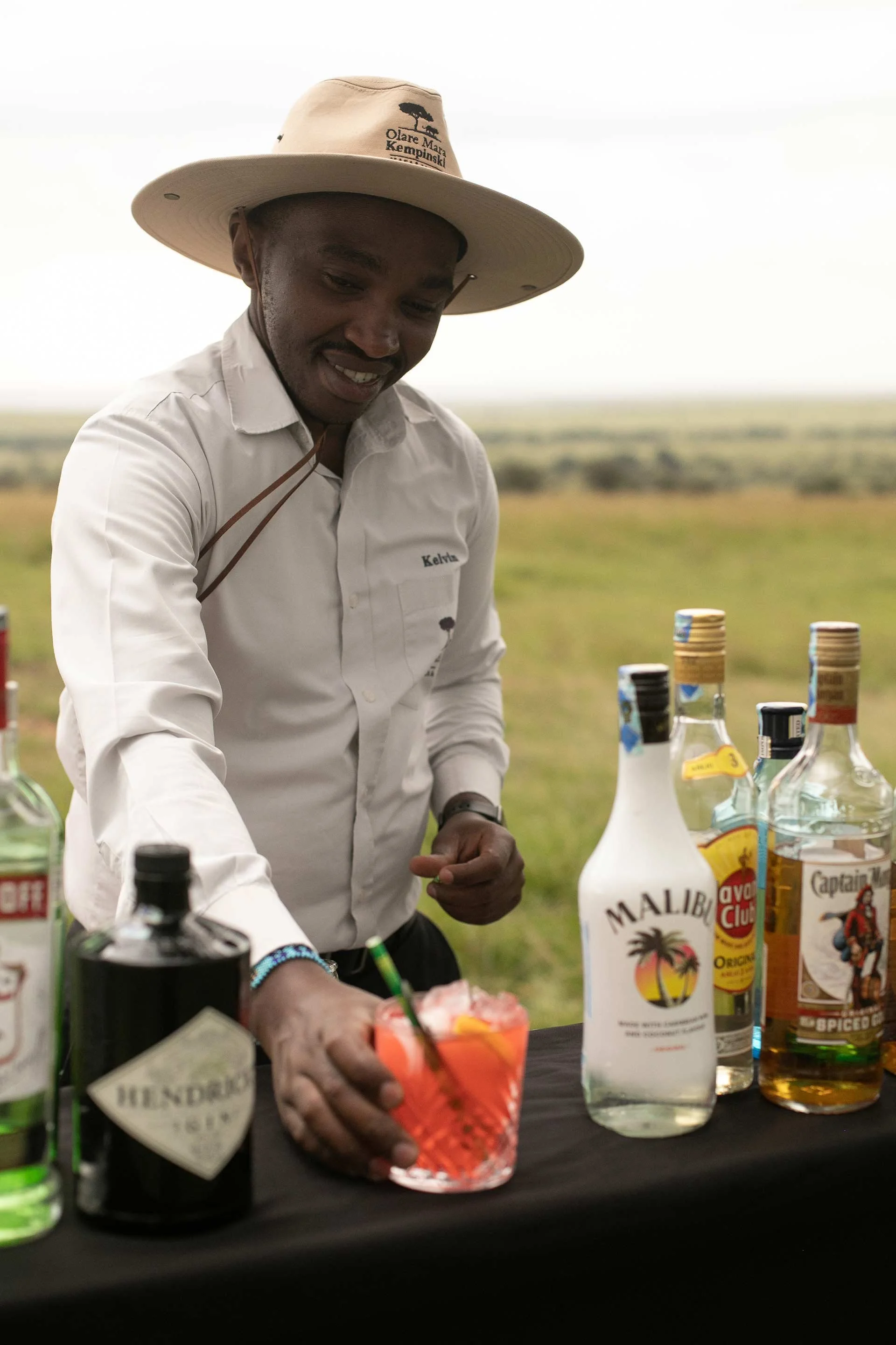 A man wearing a wide-brimmed hat and white shirt preparing a colorful cocktail at an outdoor bar with bottles of liquor, including Malibu and Captain Morgan, set against a grassy landscape.