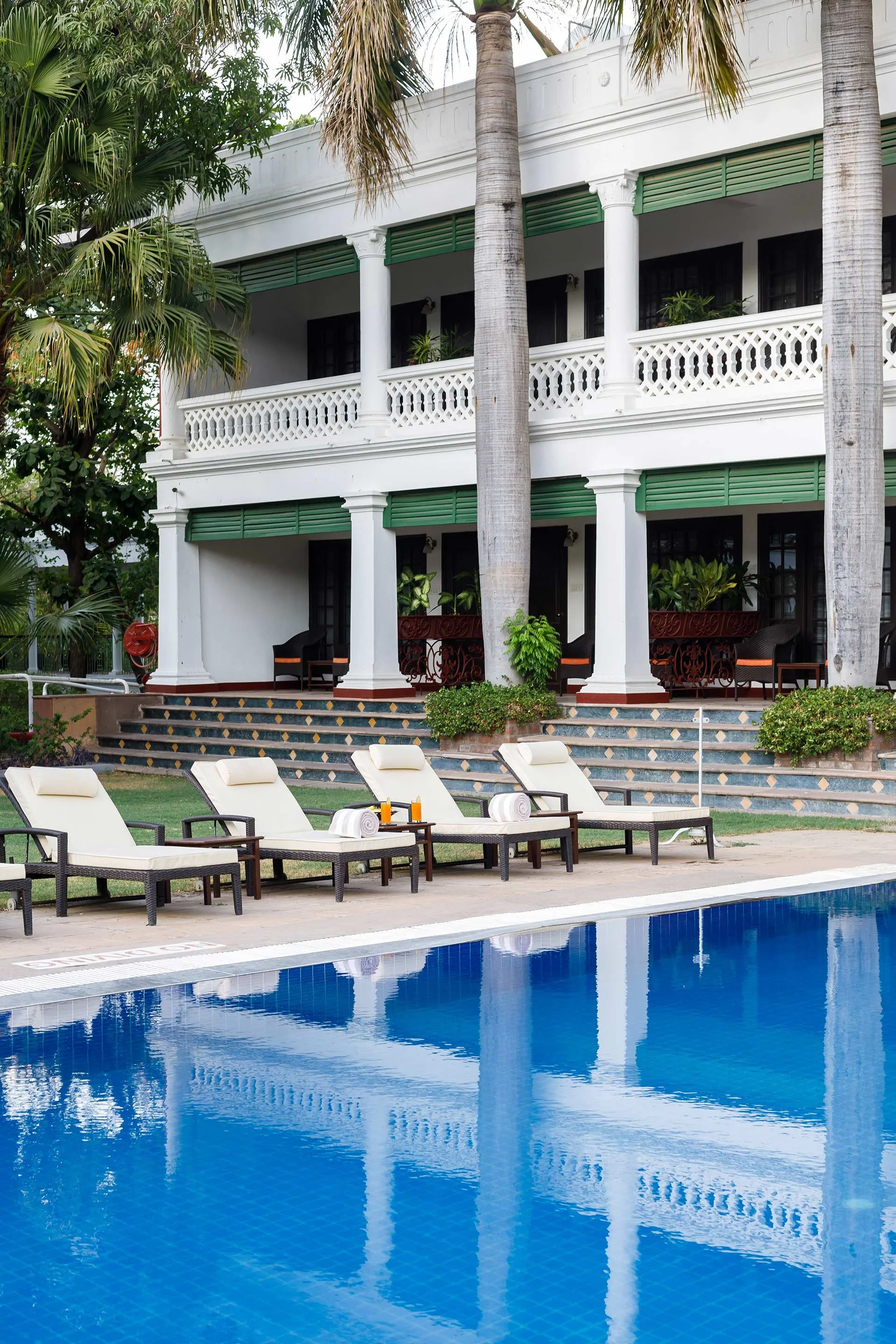 Poolside view of a white, two-story building with a balcony, surrounded by palm trees and greenery, featuring pool chairs with rolled towels and a small table with drinks.