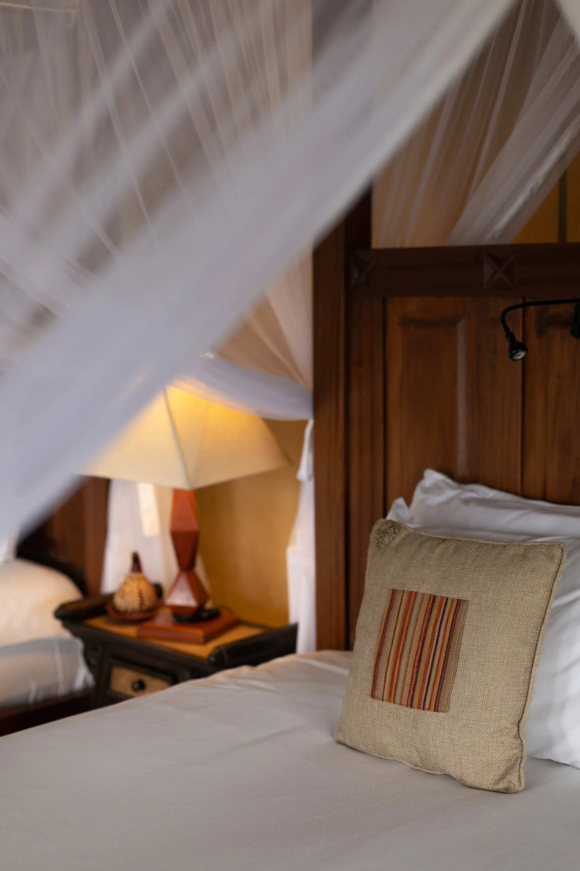 A bedroom with a bed featuring white bedding and a decorative pillow with striped pattern. A wooden headboard and side table with a lamp are visible, along with a white curtain and a portion of an umbrella in the background.
