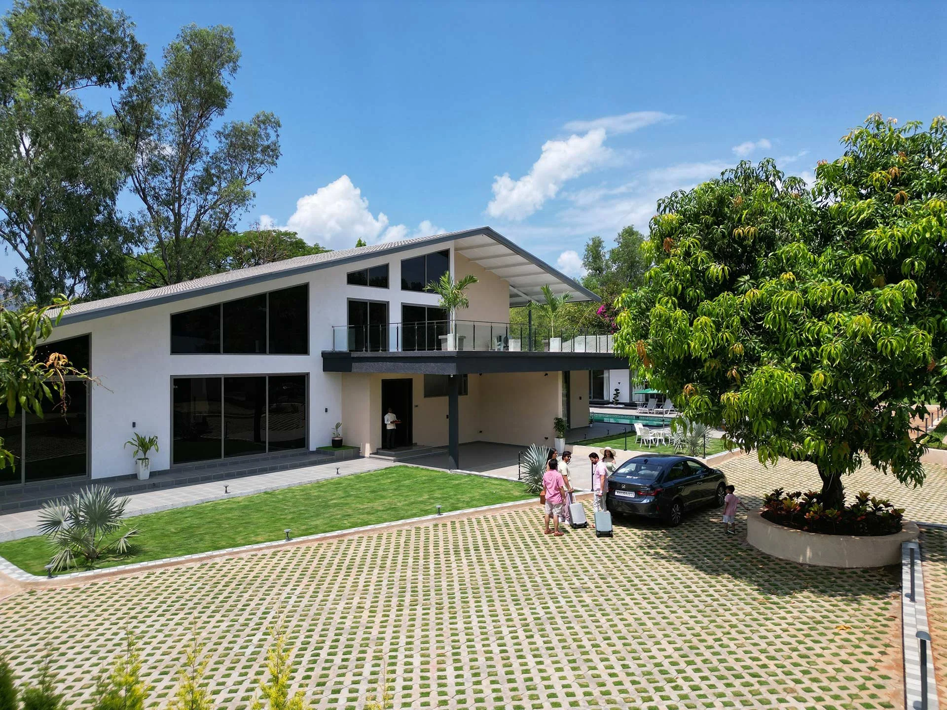 A modern house with large glass windows, a lawn, and a driveway with a tree and guests with luggage near a black car.