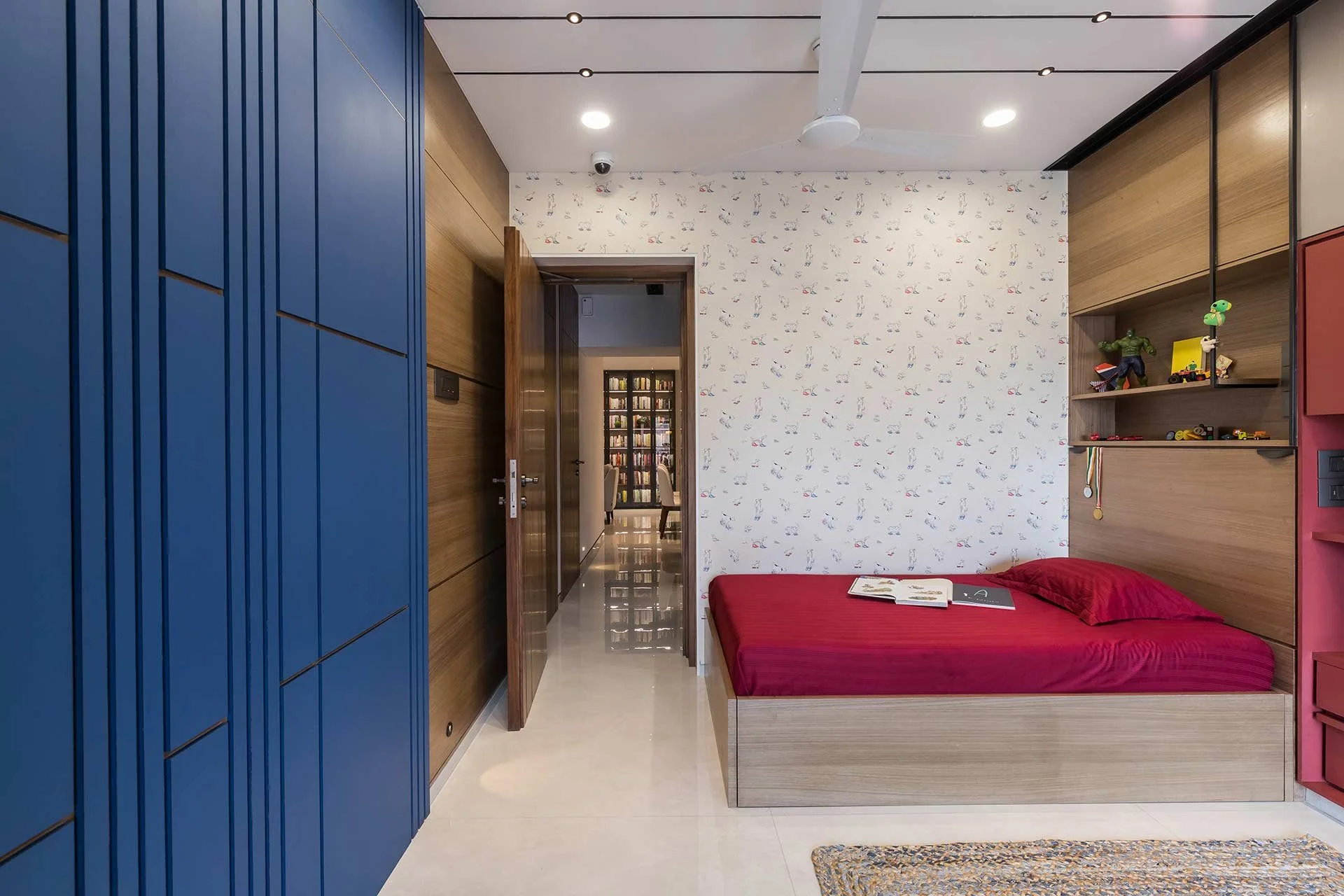 A bedroom with a bed that has a red mattress and pillow, built-in wooden shelves with toys and medals, colorful cabinets, and an open door leading to a hallway with bookshelves.