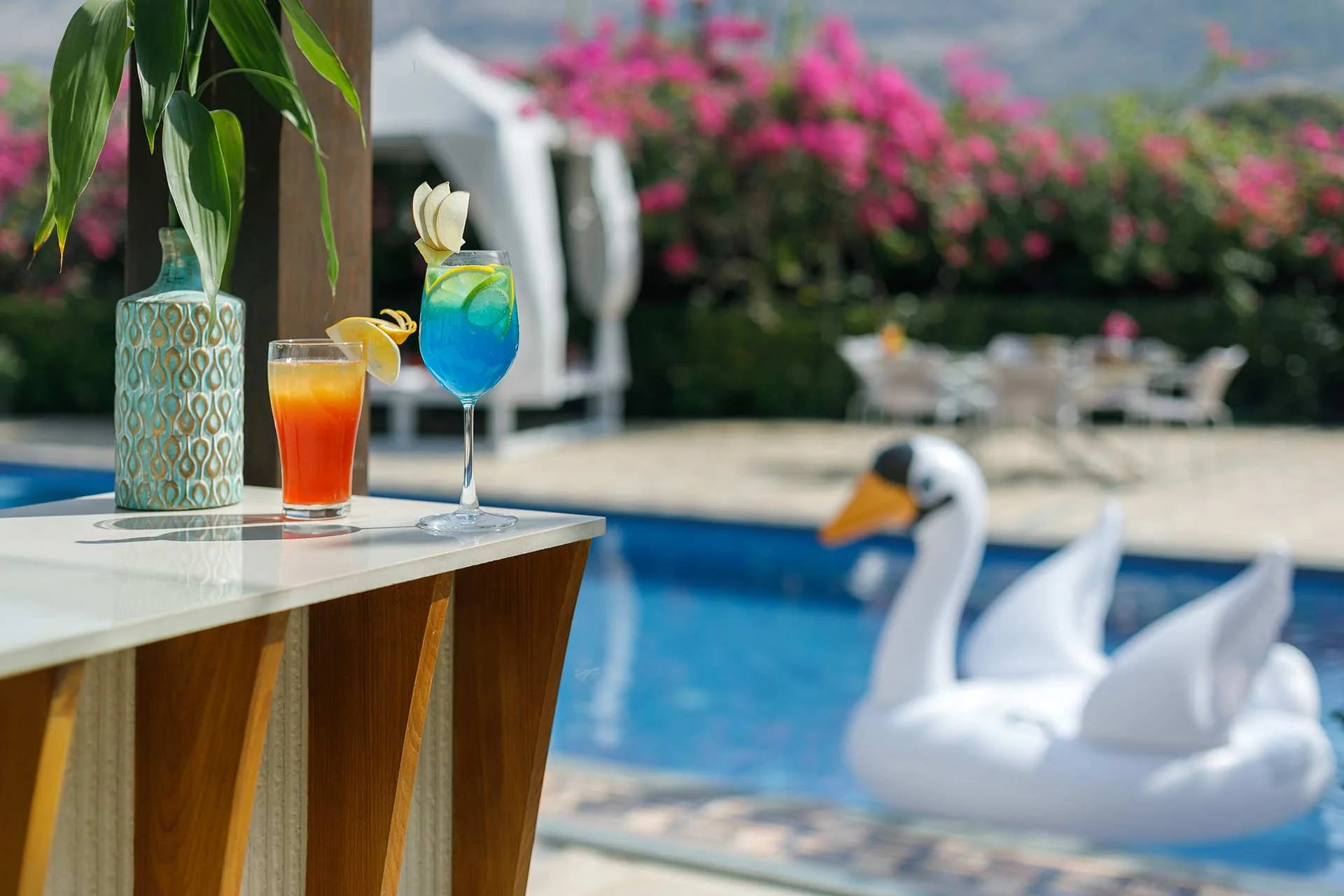 Two colorful tropical drinks on a white table near a swimming pool with white swan floats and pink flowering bushes in the background.