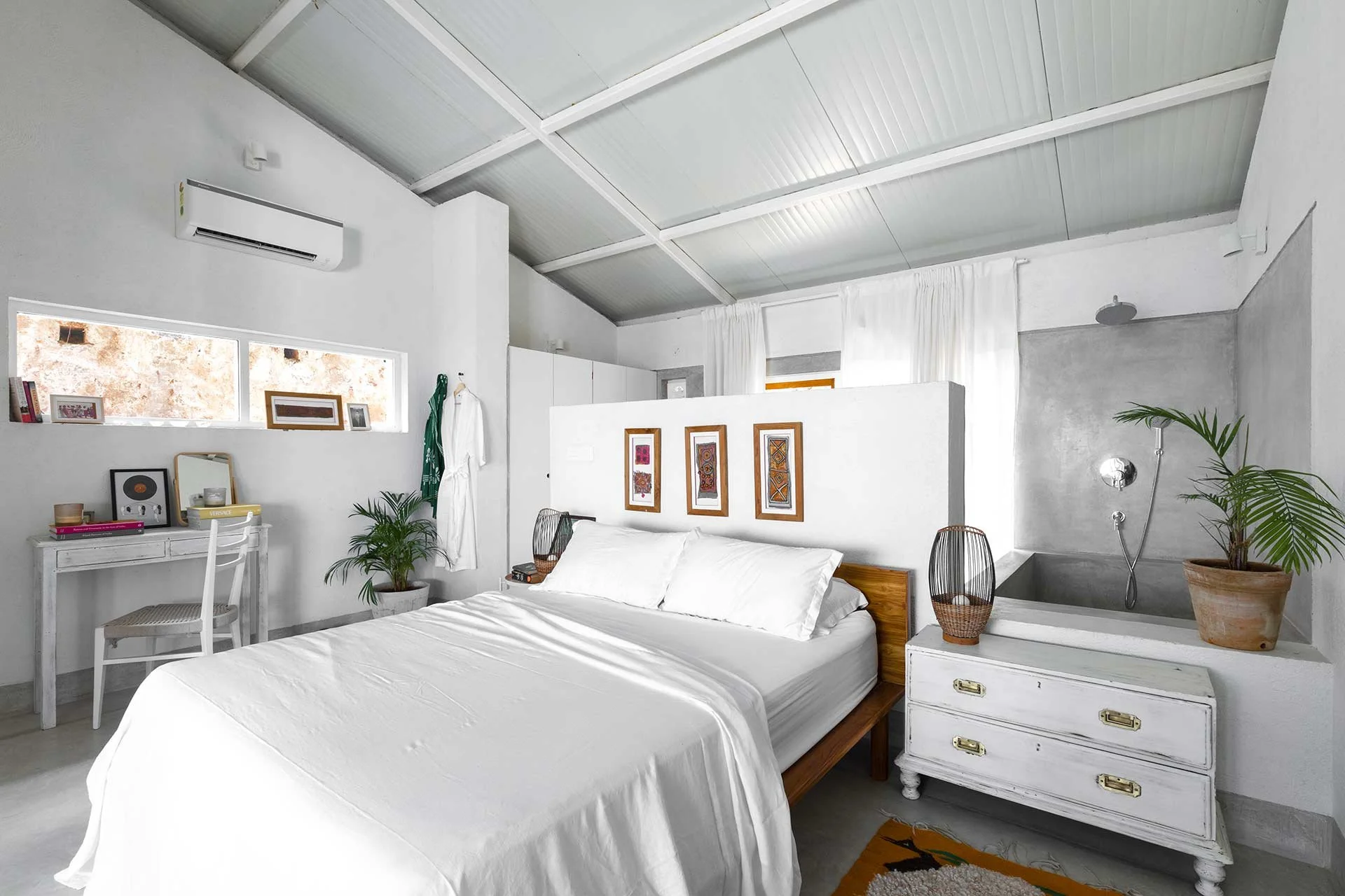 A bright, white bedroom with a mixed modern and rustic decor, featuring a white bed with pillows, a white dresser, and decorative artwork on the wall, with potted plants and a unique shower area in the corner.
