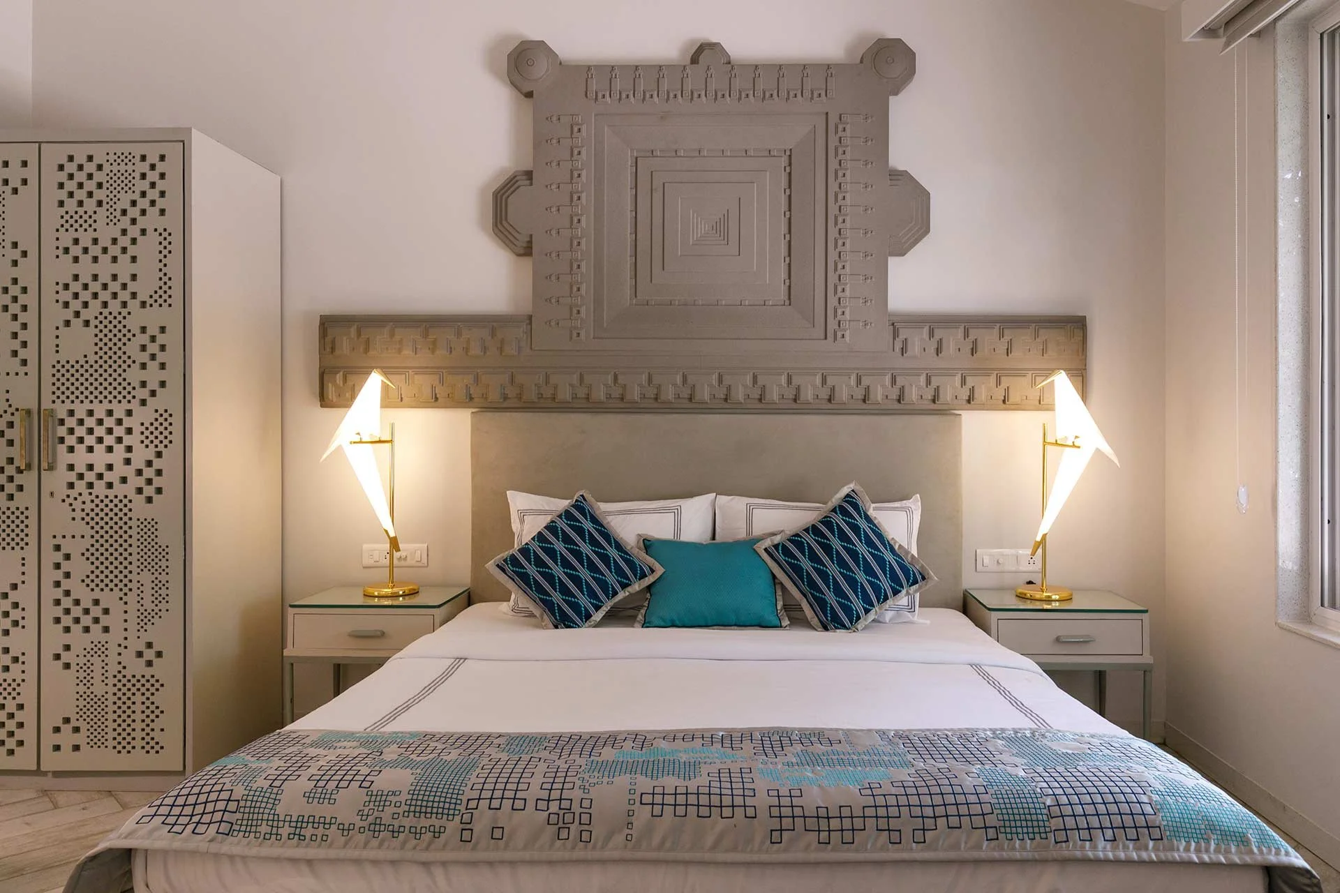 A modern bedroom with a decorative wall panel above the bed, bedside tables with gold lamps, and a bed with white linens and blue patterned pillows.