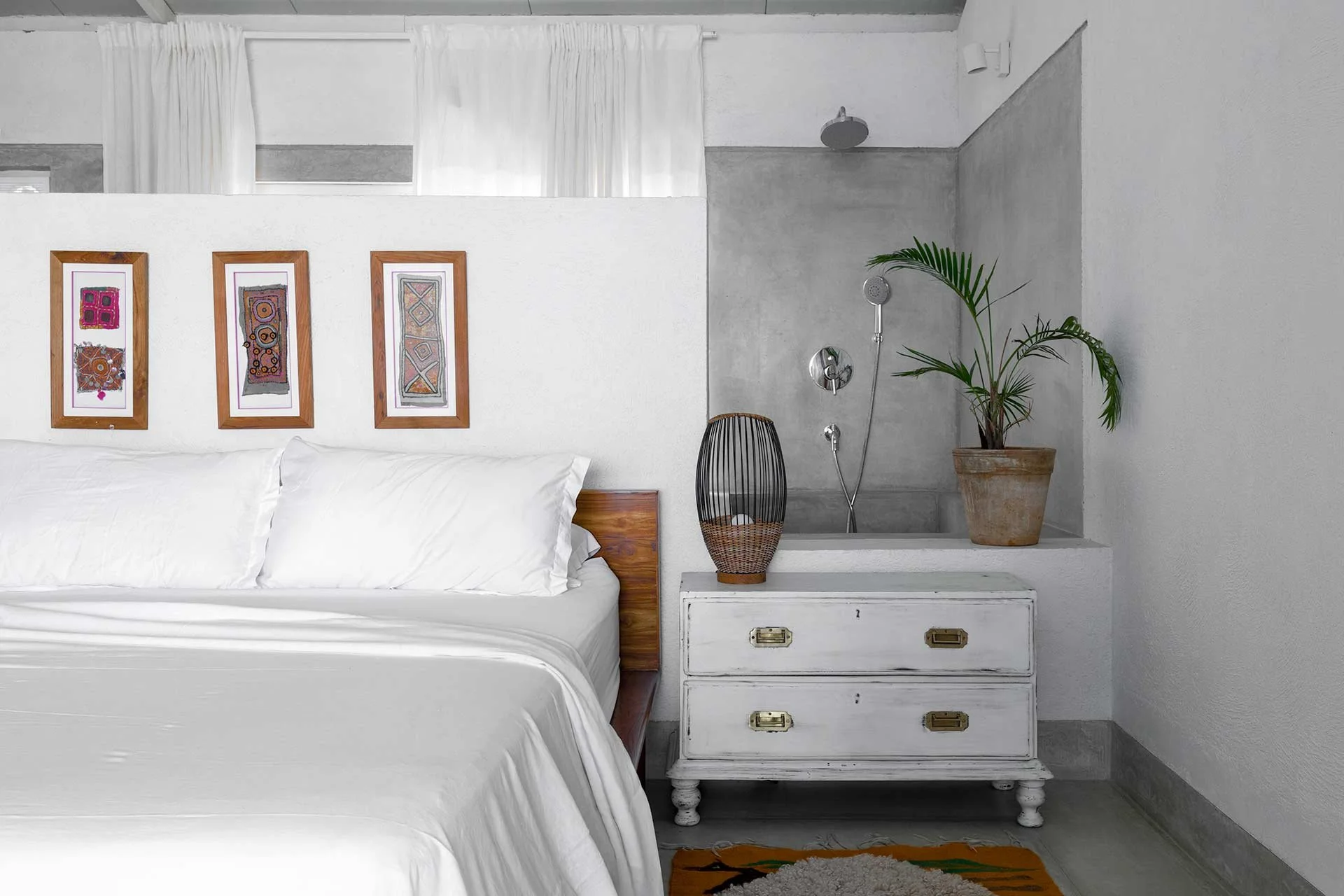 A bedroom with white walls and bedding, three framed colorful abstract art pieces above the bed, a white distressed dresser with brass handles beside the bed, topped with a woven basket, a half-plant, and a decorative lamp. A small shower area with a