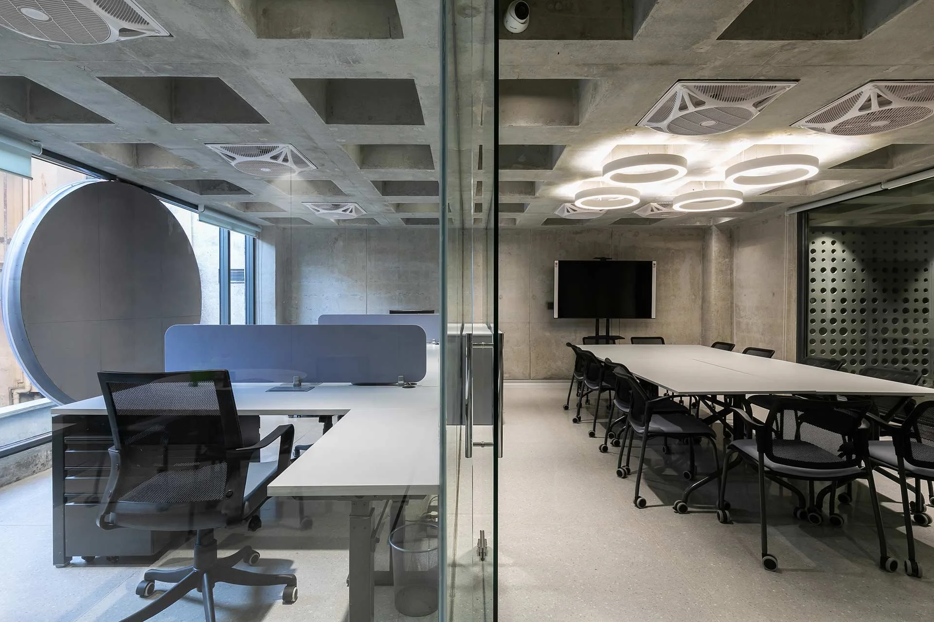 Modern office conference room with glass wall dividing two parts; one side has a desk with an office chair, the other side has a long table with multiple chairs and a TV mounted on the wall.