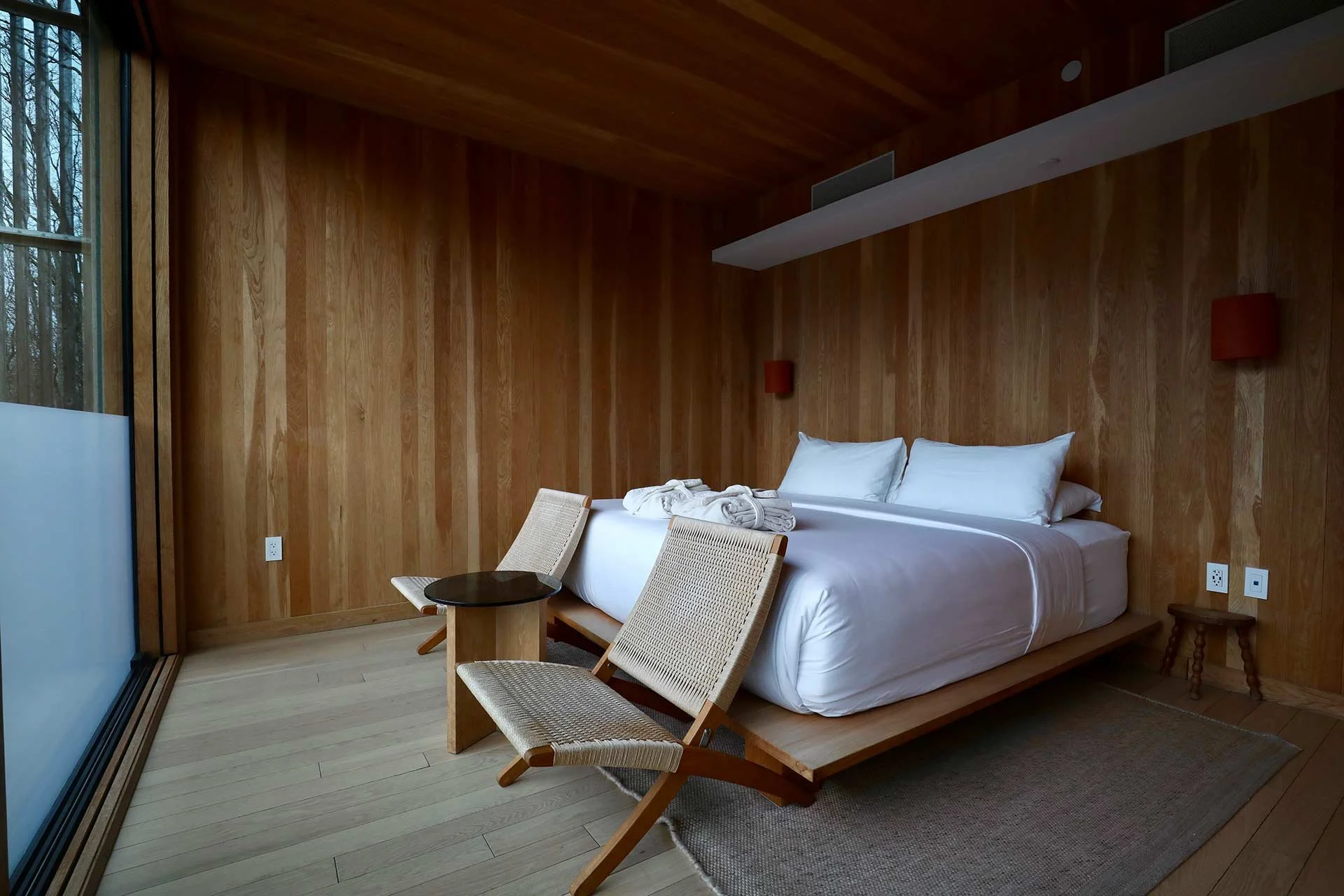 Room with wooden walls and ceiling, large bed with white linens and pillows, two foldable chairs, small round black table, large glass window showing trees outside, two wall-mounted lamps, small wooden side table, and electrical outlets on the wall.