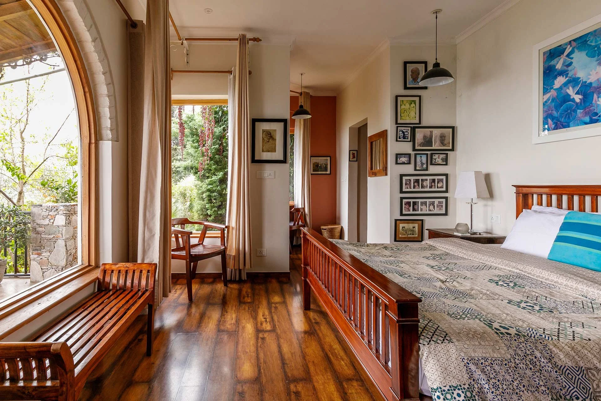 A cozy bedroom with a large window showing a view of a garden. The room contains a wooden bed with patterned bedding, a nightstand with a lamp, and chairs near the window. The walls are decorated with framed photos and artwork.