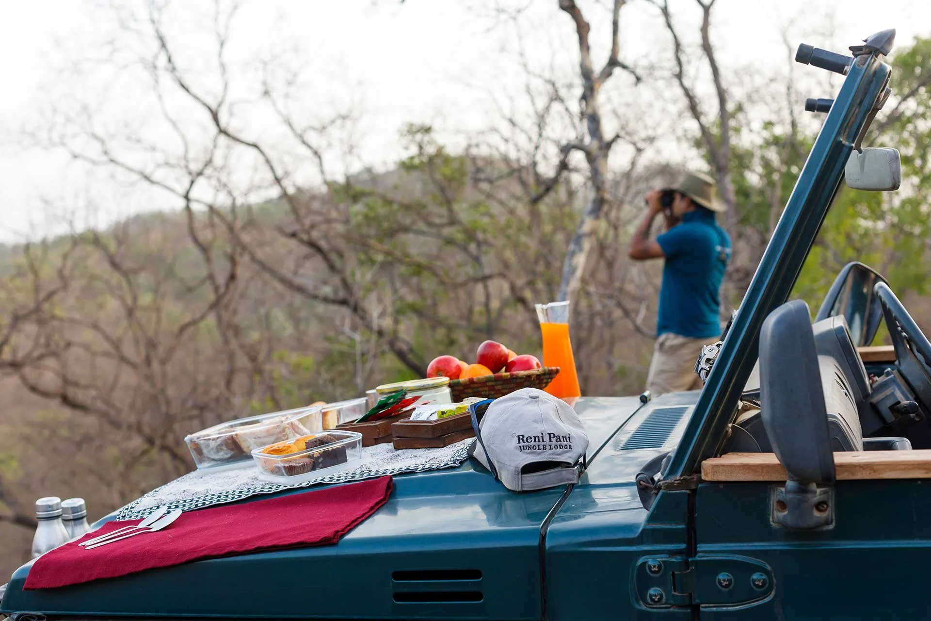 A picnic setup on the hood of a green off-road vehicle, with food, drink, and utensils, while a man in a blue shirt and hat is looking into the distance through binoculars, surrounded by a background of leafless trees.
