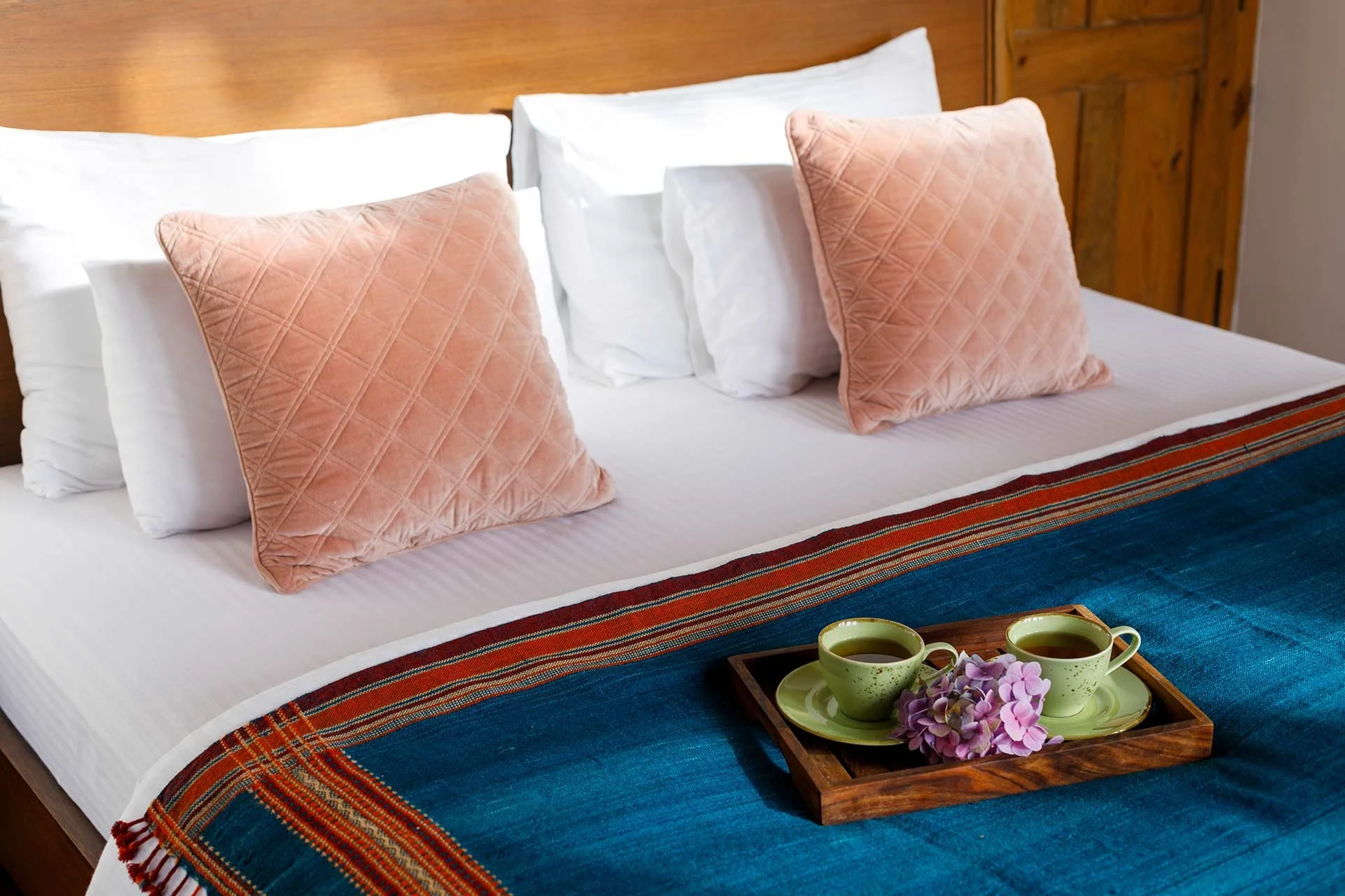 A neatly made bed with white sheets, four white pillows, and two peach-colored quilted pillows. A wooden headboard is visible, and a tray with two cups of tea and purple flowers rests on a blue and multicolored bedspread. In Nainital Uttarakhand.