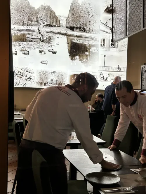 Zwei Köche in einem Restaurant bereiten einen Tisch vor, im Hintergrund ein großes Fenster mit Blick auf eine Stadt mit Wasser und Bäumen.