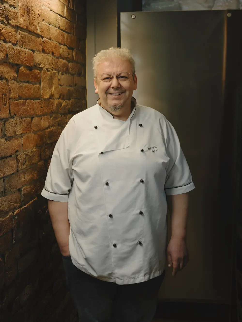 Chefkoch steht vor einer Backsteinmauer hinter dem Holzkohlengrill und lächelt.